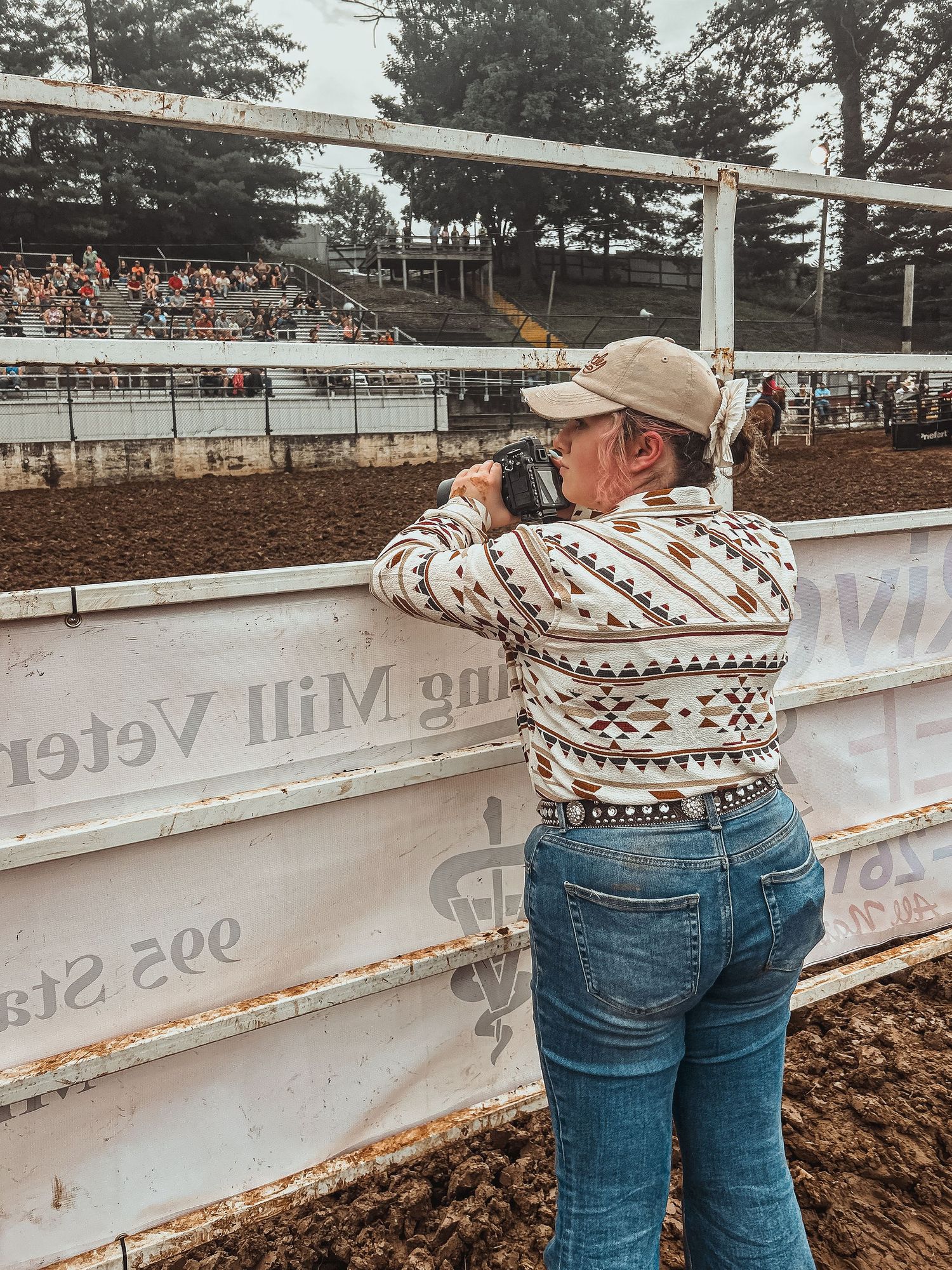 Woman in patterned sweater and jeans photographing a rodeo arena from behind white fence rails.