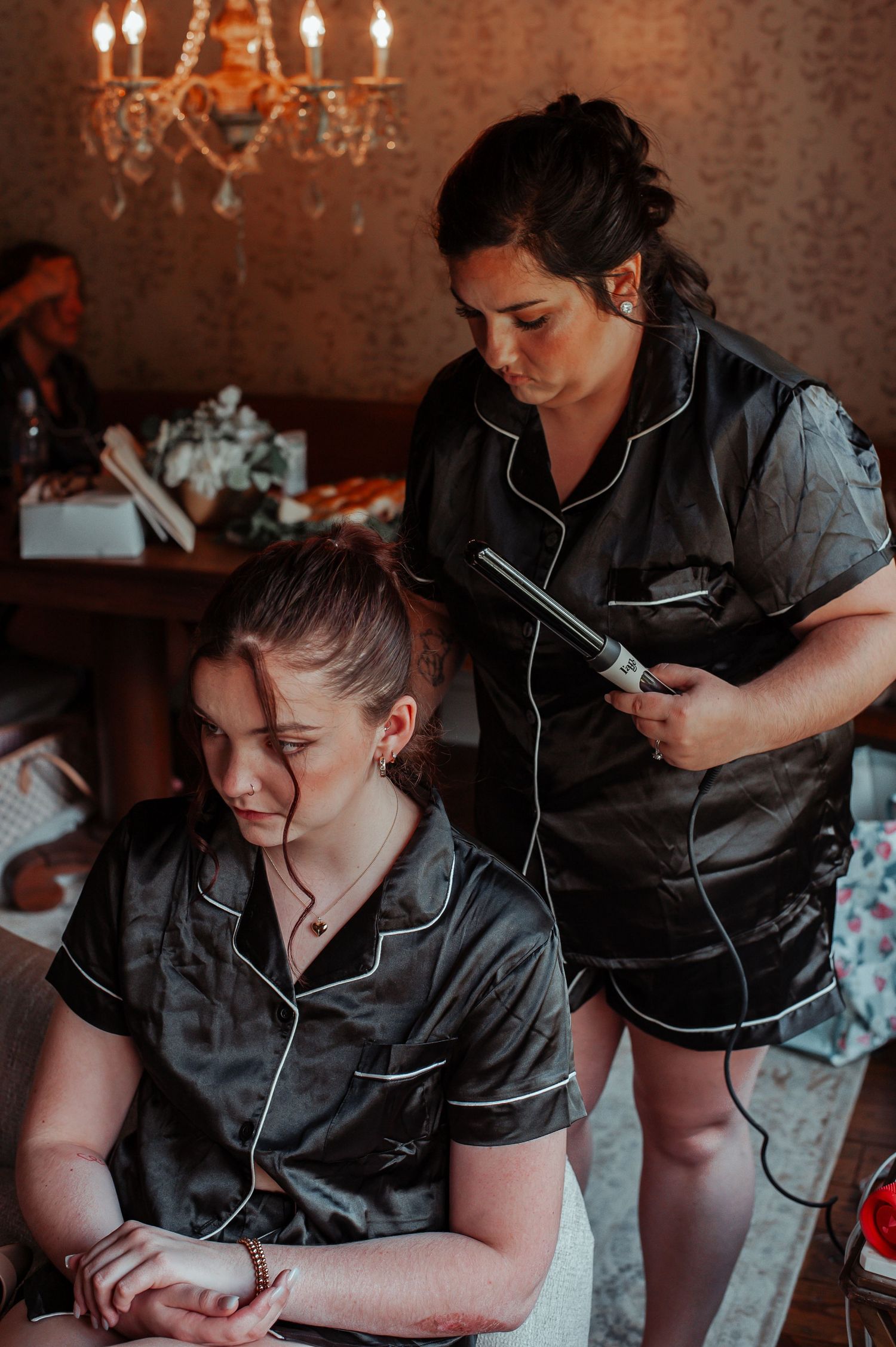 Bridesmaid in black satin pajamas gets hair styled with curling iron under elegant chandelier.