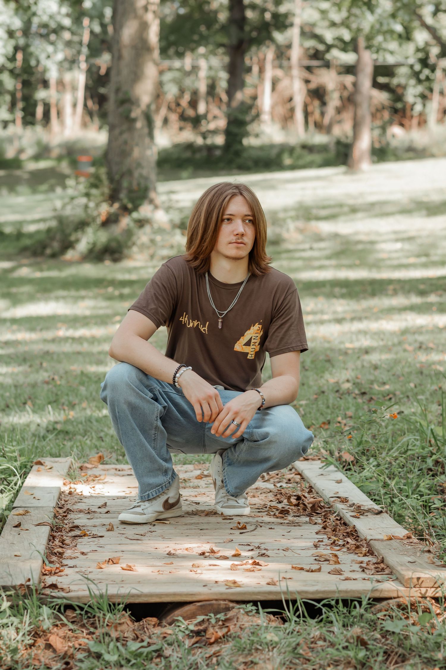 Young man with long hair crouches on wooden boards in park wearing brown graphic tee and jeans.