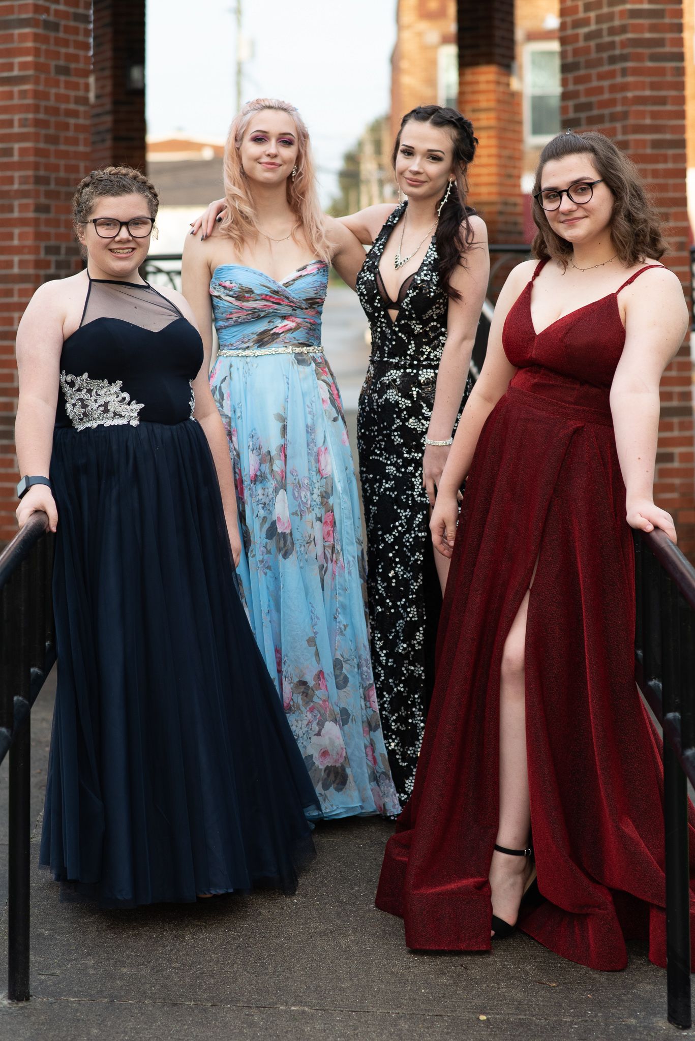Four young women in colorful formal prom dresses posing together on outdoor brick staircase.