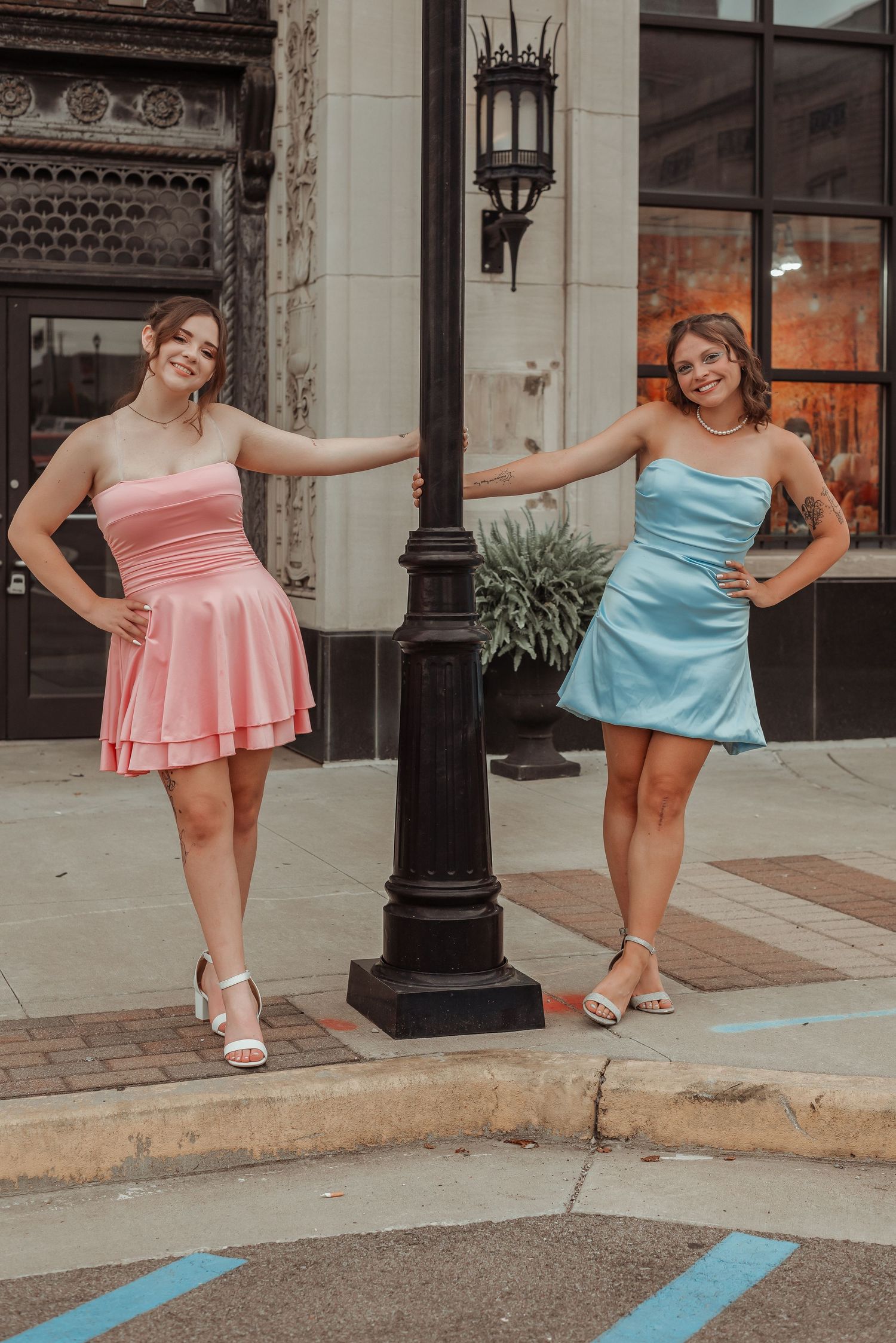 Two women in pink and blue strapless dresses posing with a black street lamp.