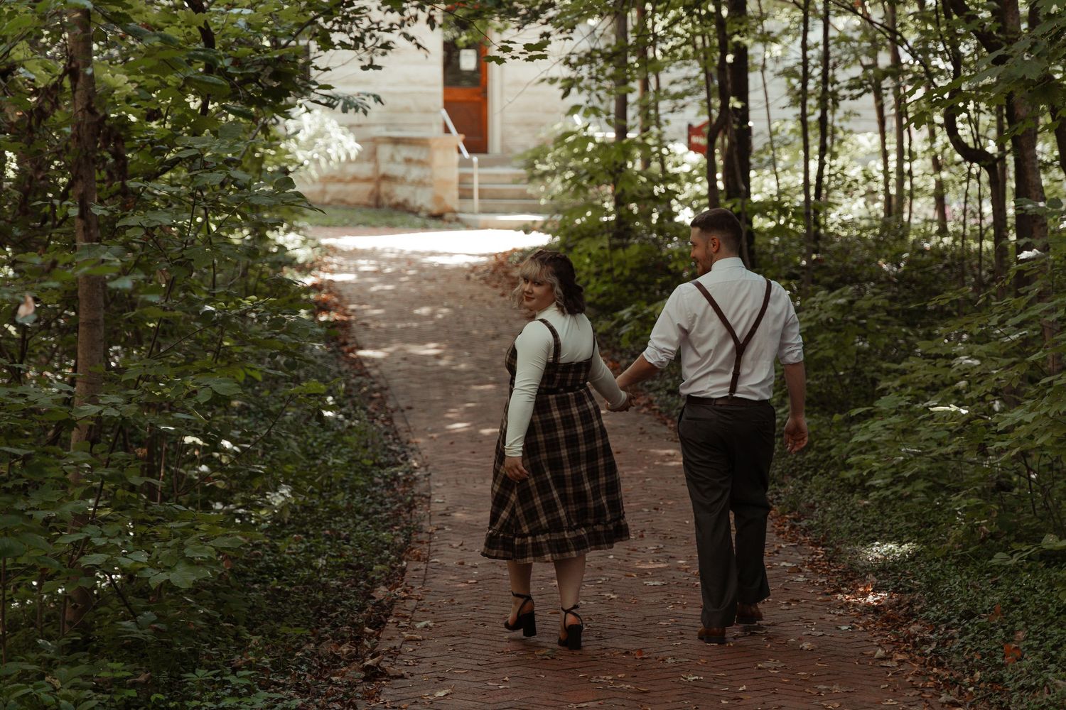 Couple walking hand in hand down a tree-lined brick pathway, woman in plaid dress and man in suspenders.