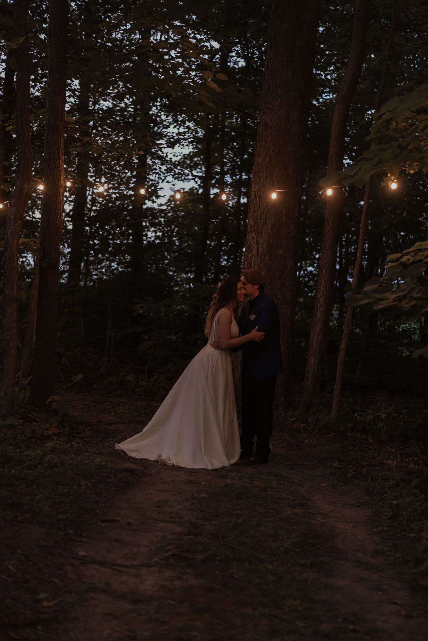 Romantic couple embraces against a tree on a forest path with warm string lights overhead.