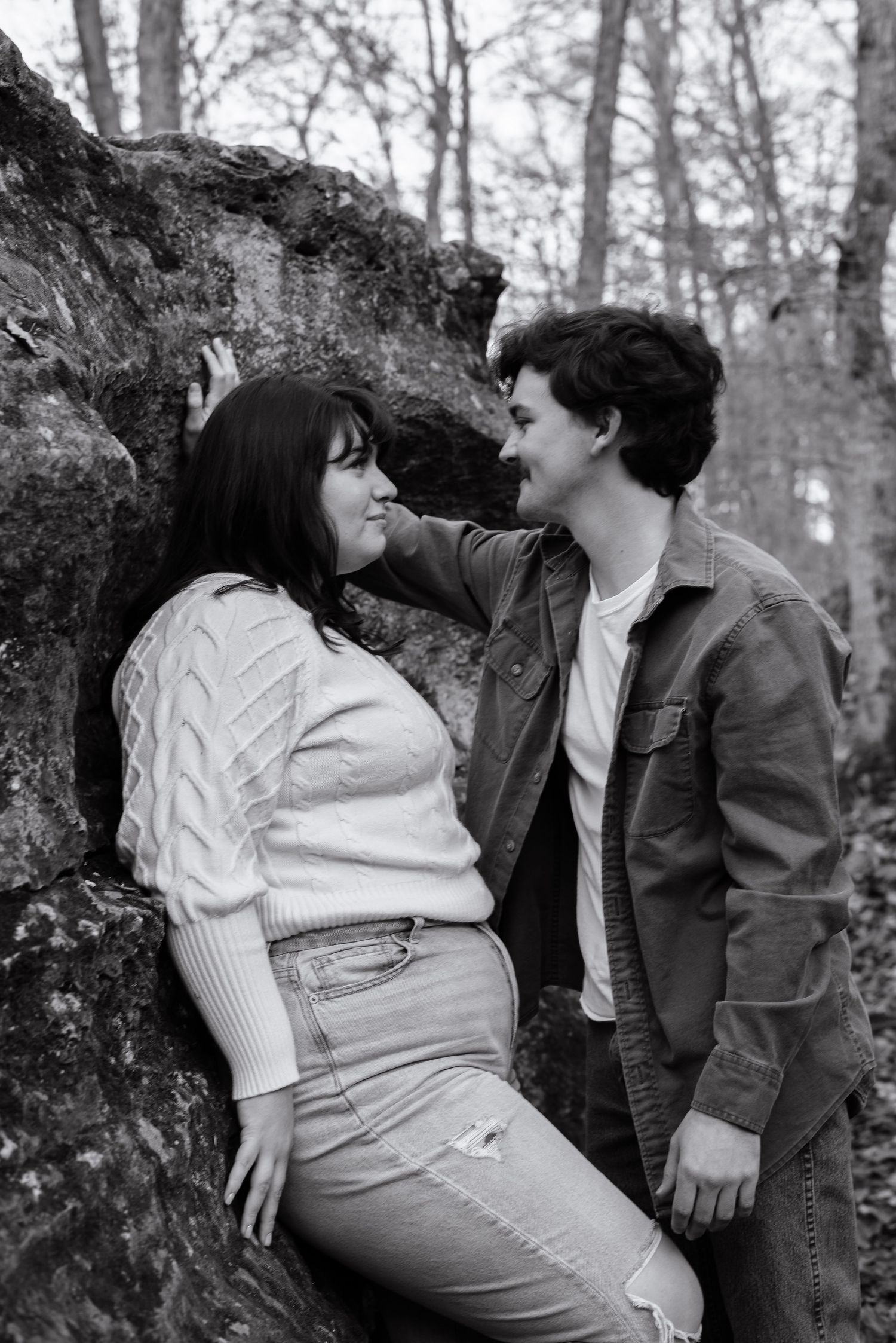 A black and white romantic photo of a couple leaning against a mossy rock in the woods.