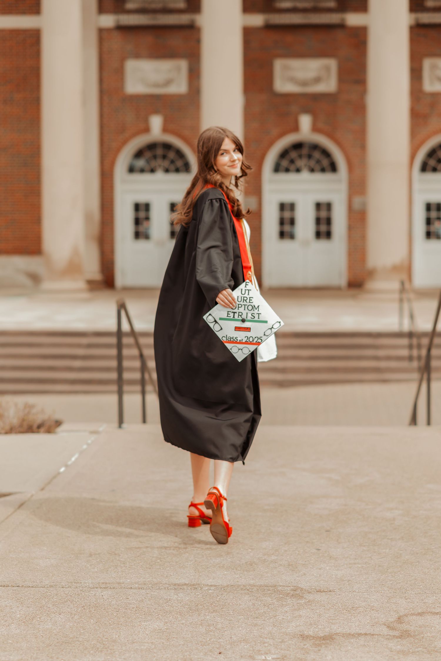 Joyful graduate in black commencement gown twirling outside a brick university building in red heels.