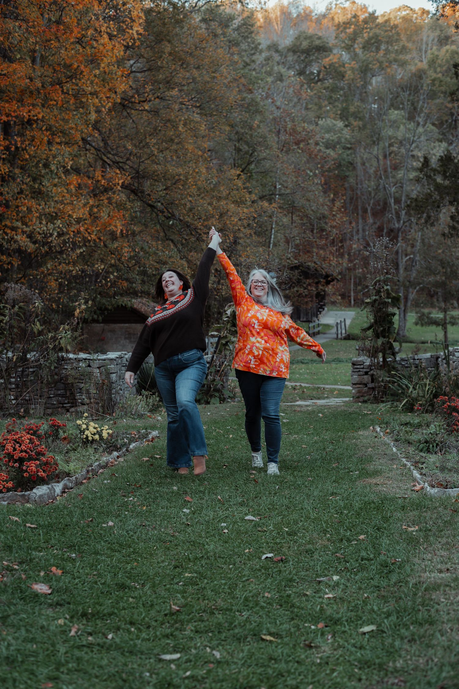 Two women joyfully dancing and holding hands in an autumn garden path.
