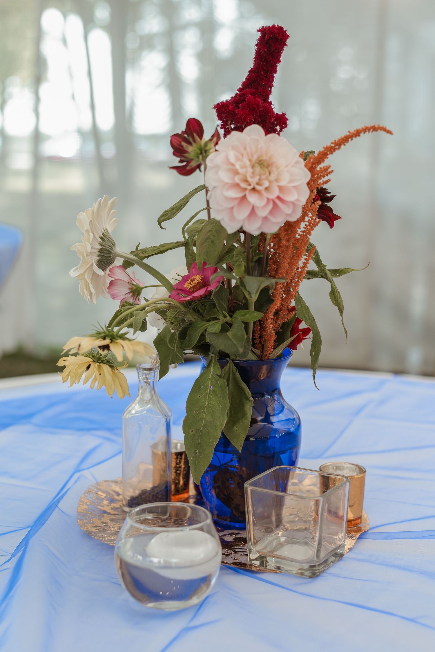Blue vase centerpiece with dahlias, amaranth, and wildflowers on light blue tablecloth with candles.
