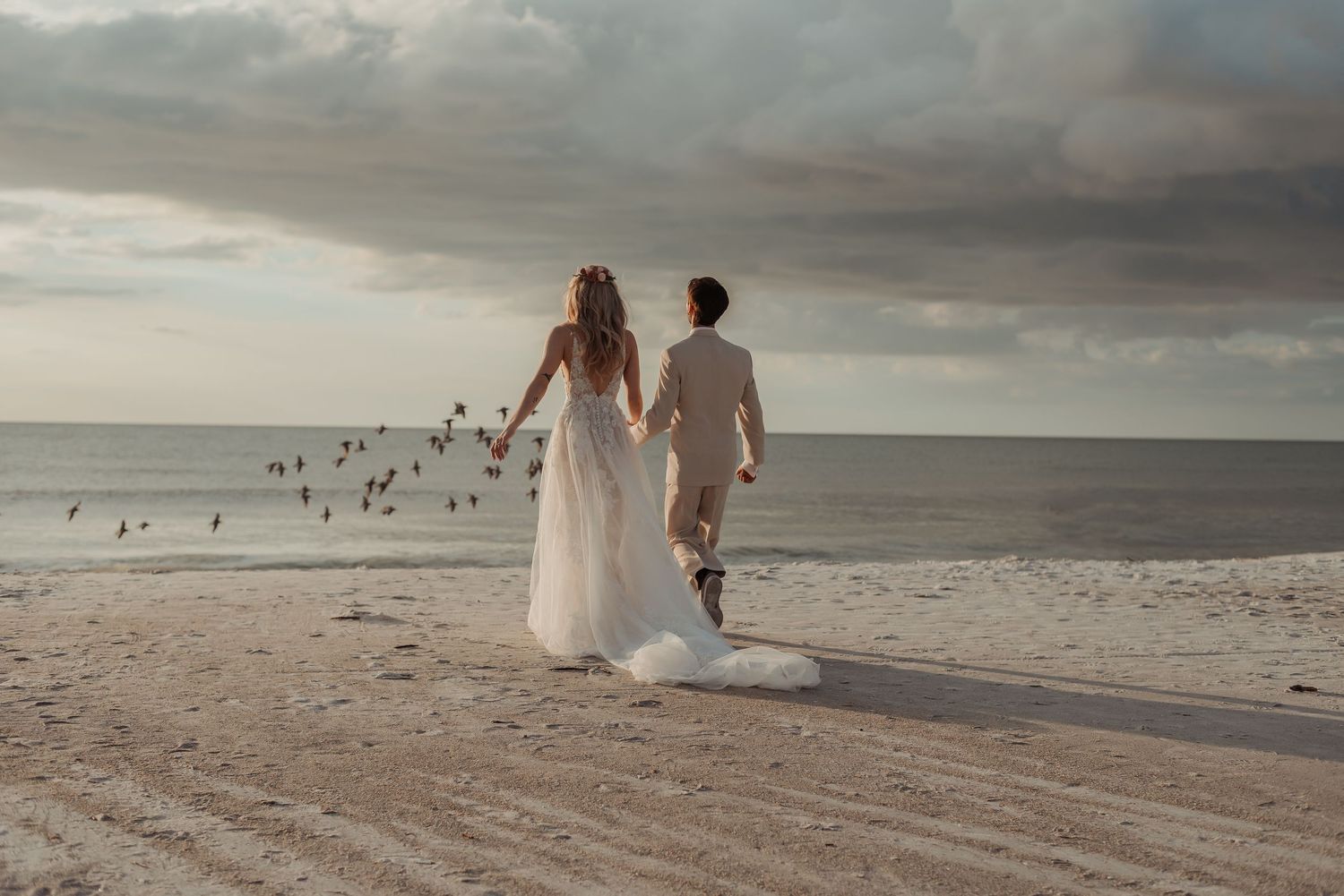 Bride and groom walking hand in hand on a sandy beach at sunset with birds flying in the background.