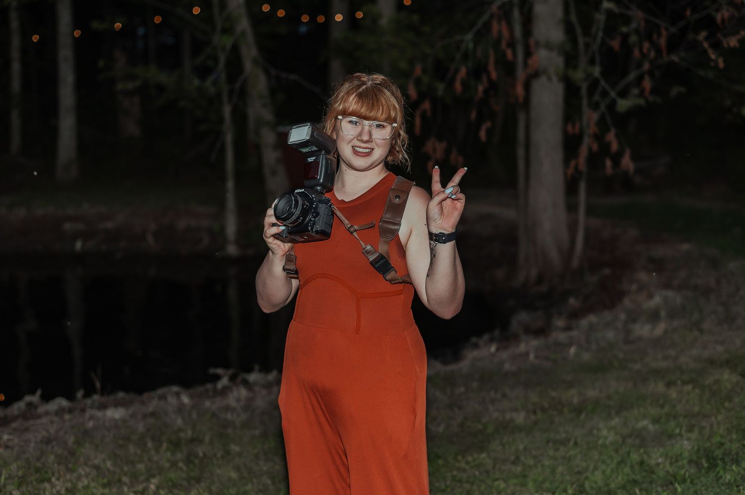 Woman in red jumpsuit holding camera and making peace sign outdoors at night.