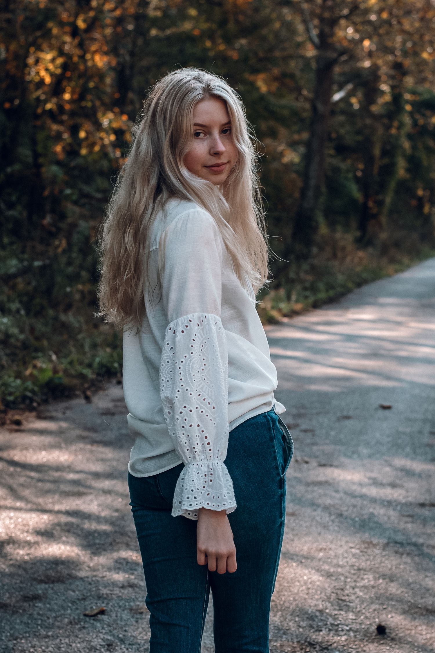 Young blonde woman in white eyelet blouse and teal pants stands on tree-lined path looking back.