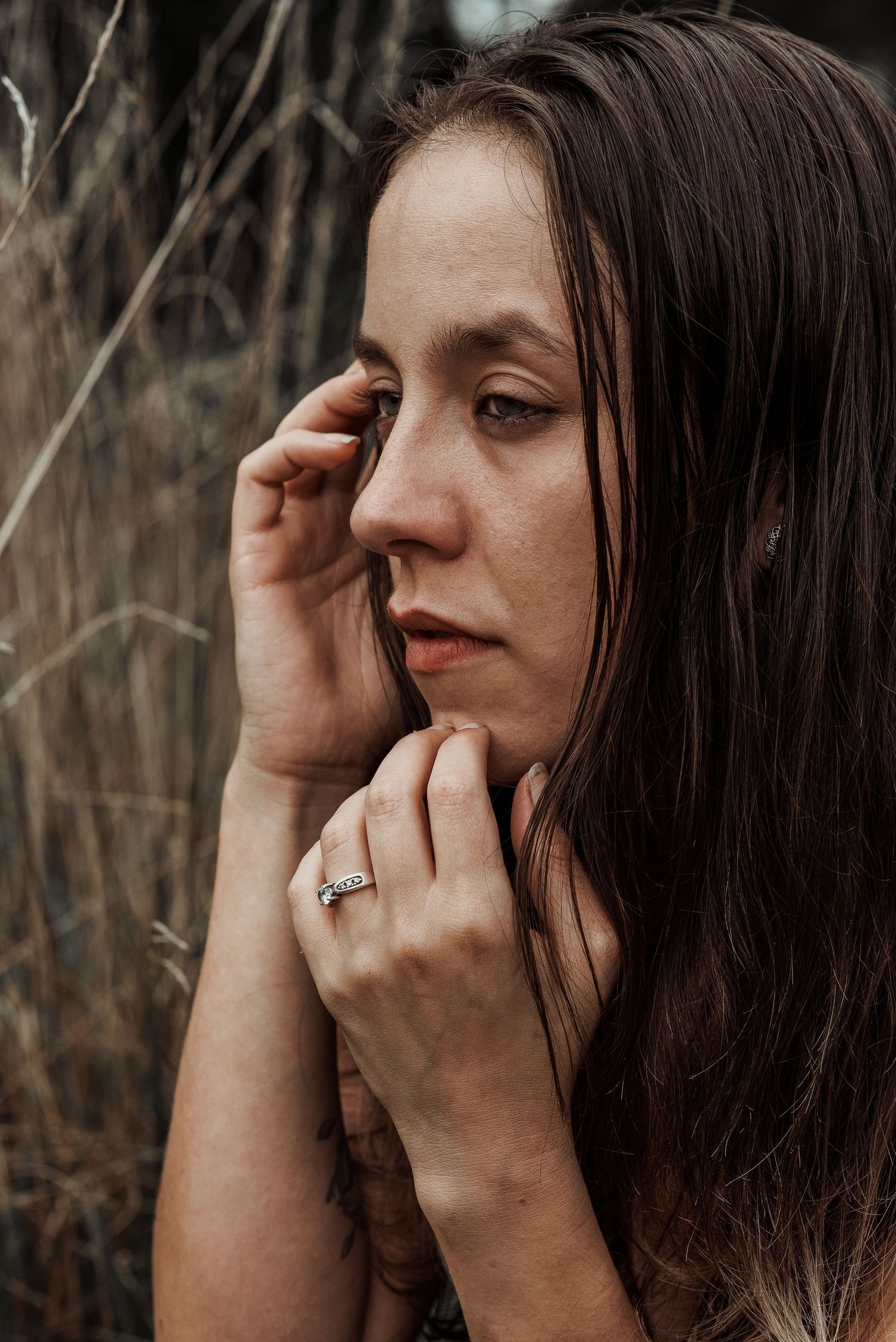 Close-up portrait of brunette woman with rings touching her face near dry grass.