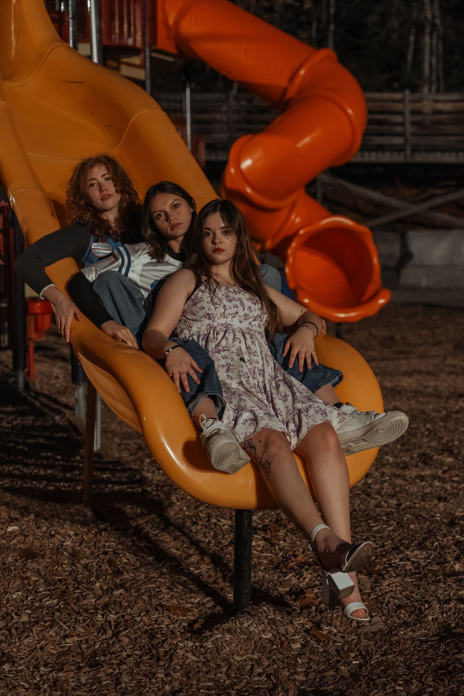 Three young women posing together on yellow playground slide at night.