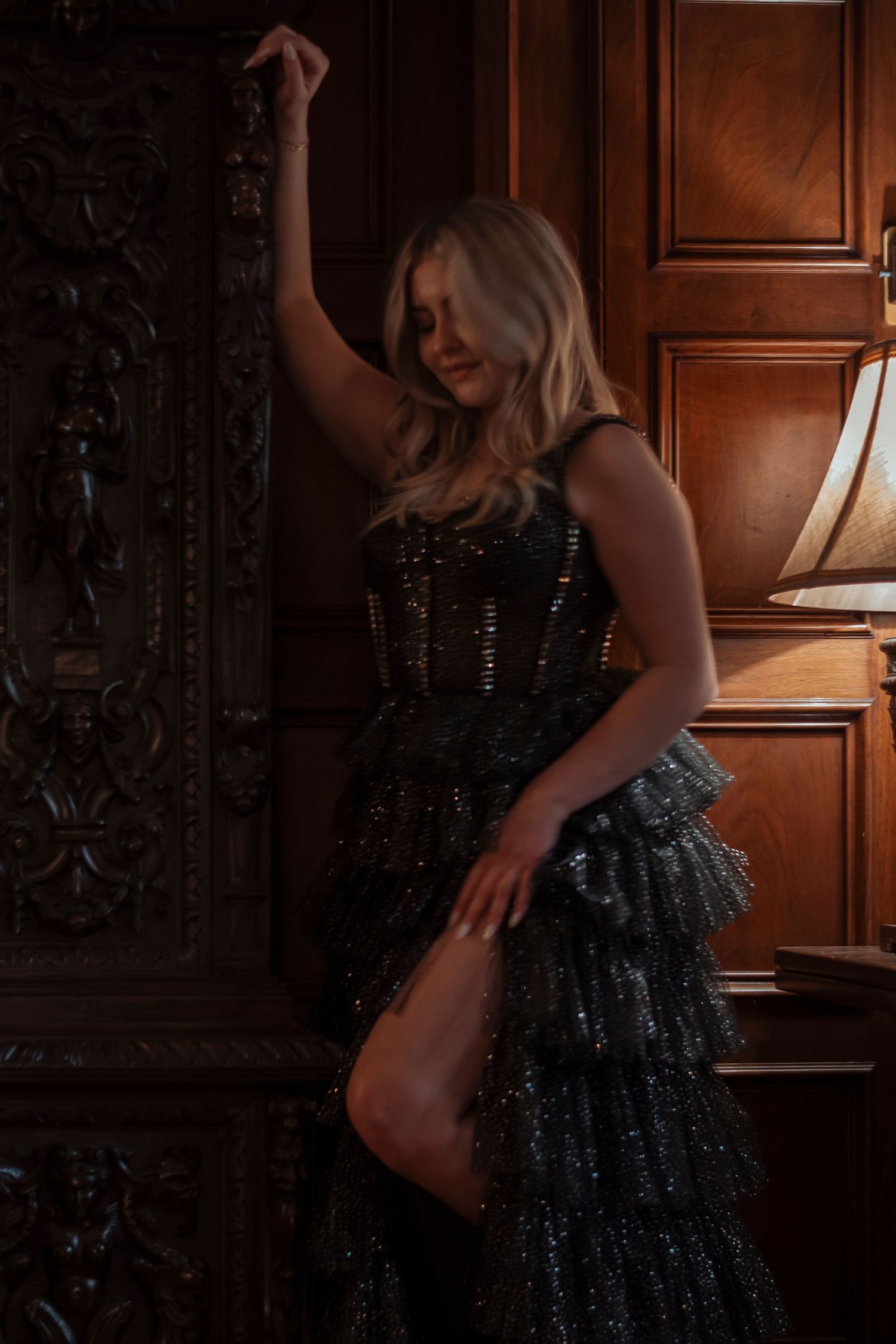 Blonde woman in sparkly dark gown leaning against carved wooden cabinet in dimly lit room.