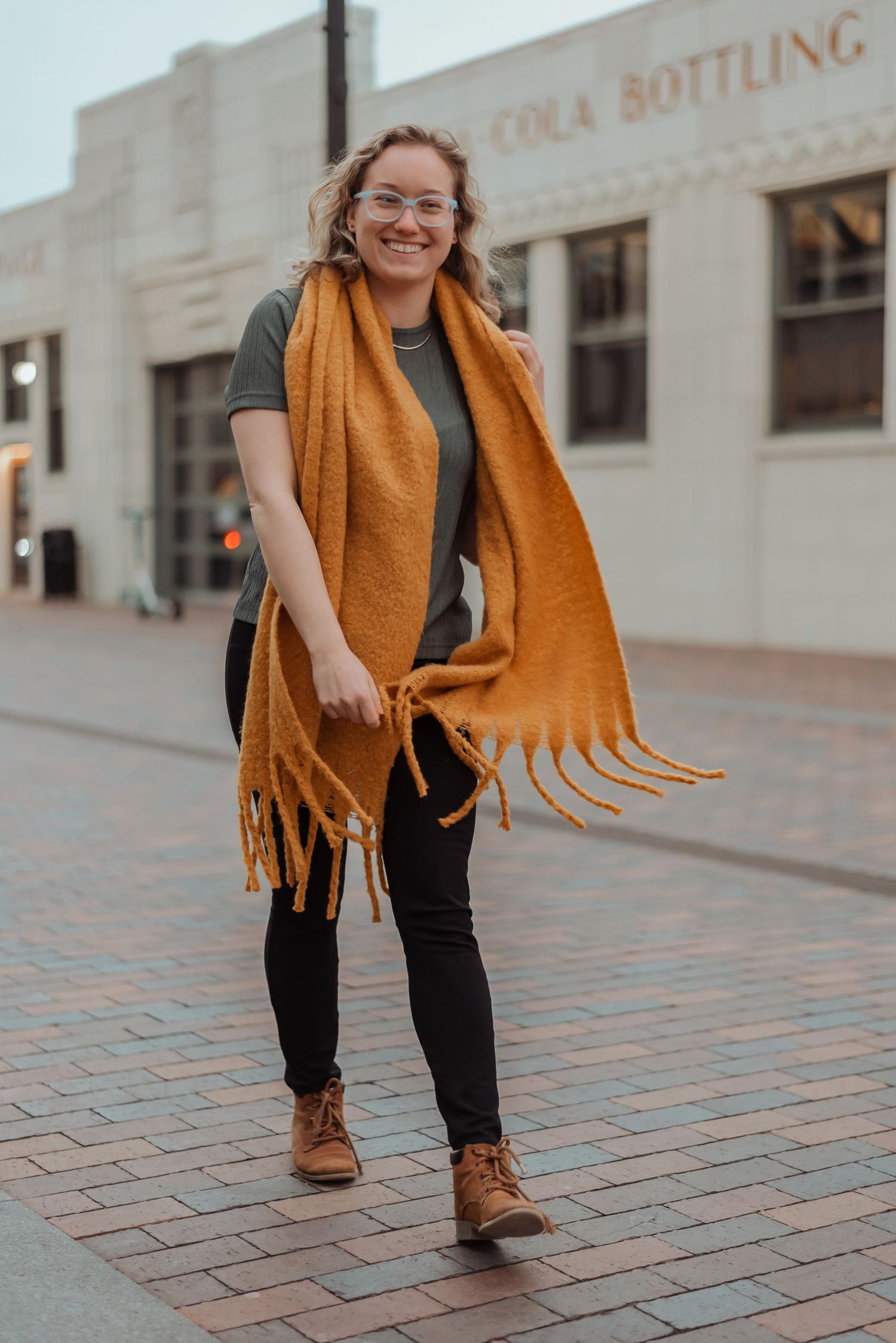 Cheerful young woman in mustard fringe scarf and brown boots walking on brick plaza near bottling building.