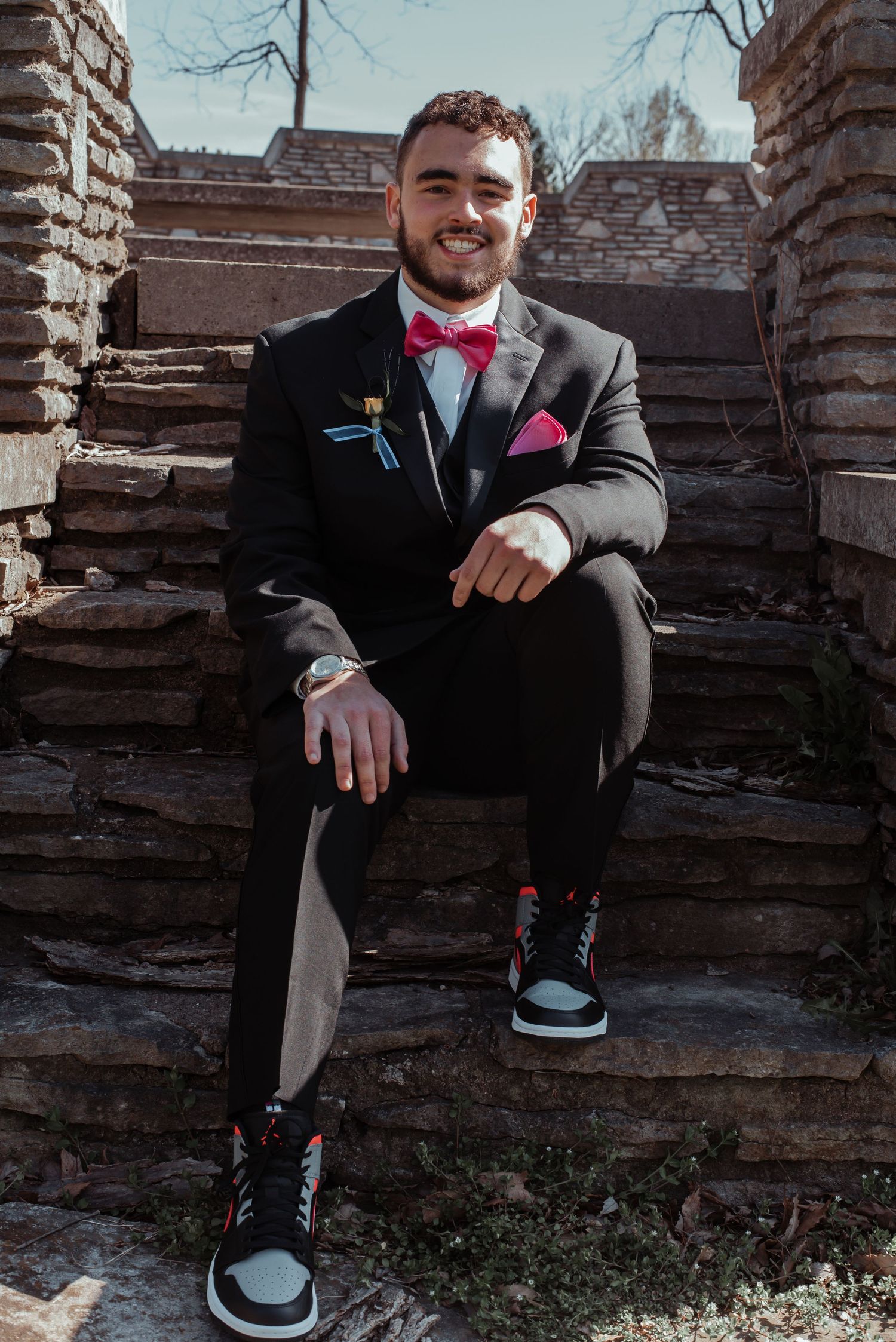 Young man in black tuxedo with red bow tie and Jordan sneakers sitting on stone steps smiling.