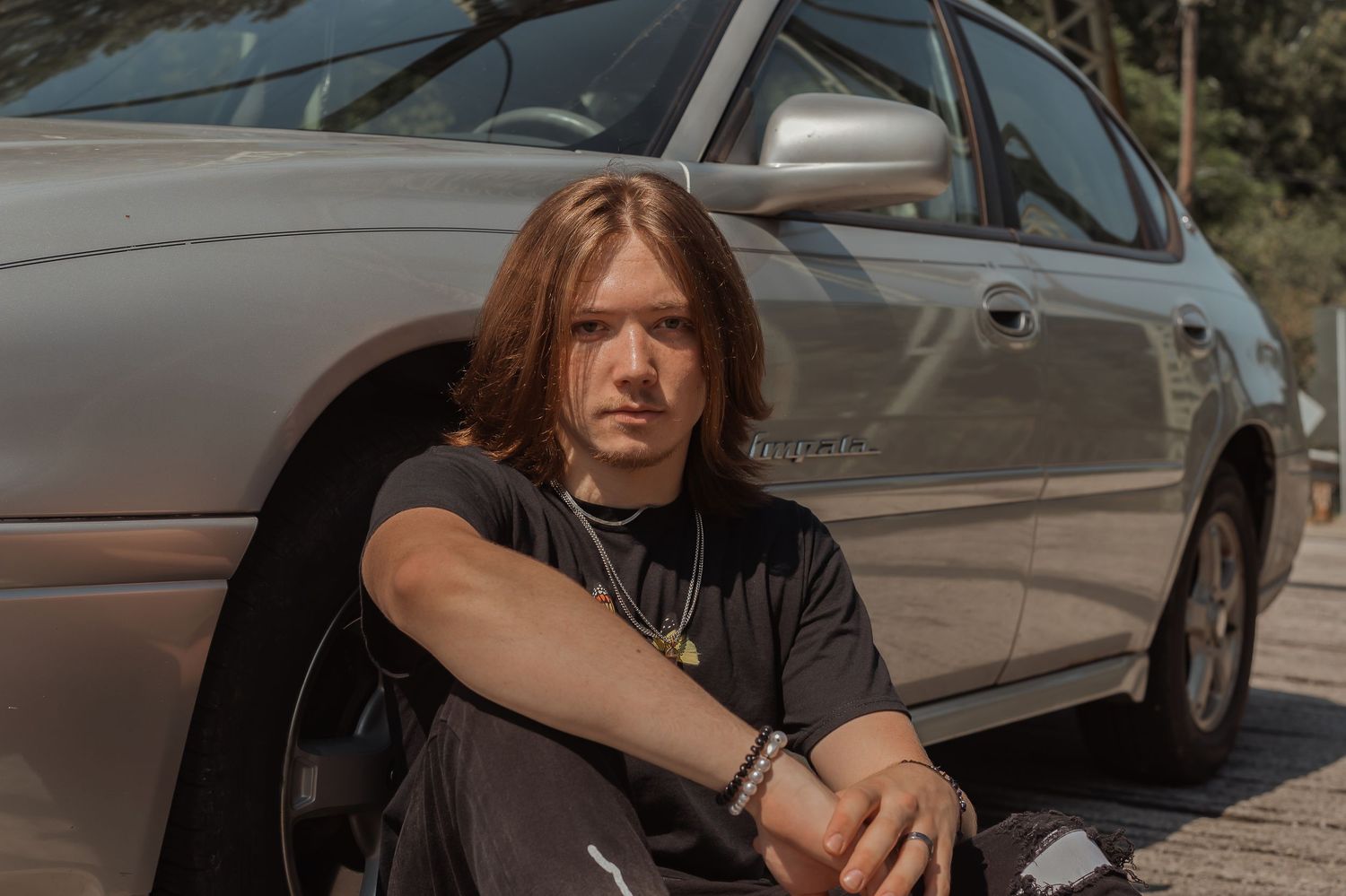 Young man with long brown hair sits against silver car tire wearing black outfit outdoors.