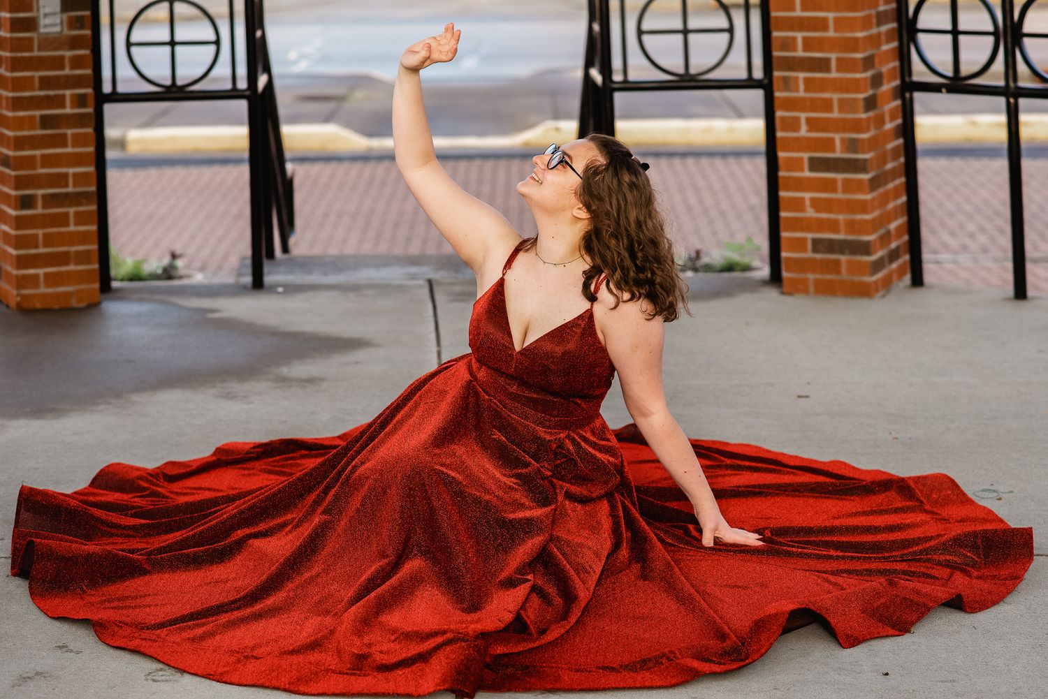 Woman in elegant red velvet ball gown posing dramatically on ground outside brick building.