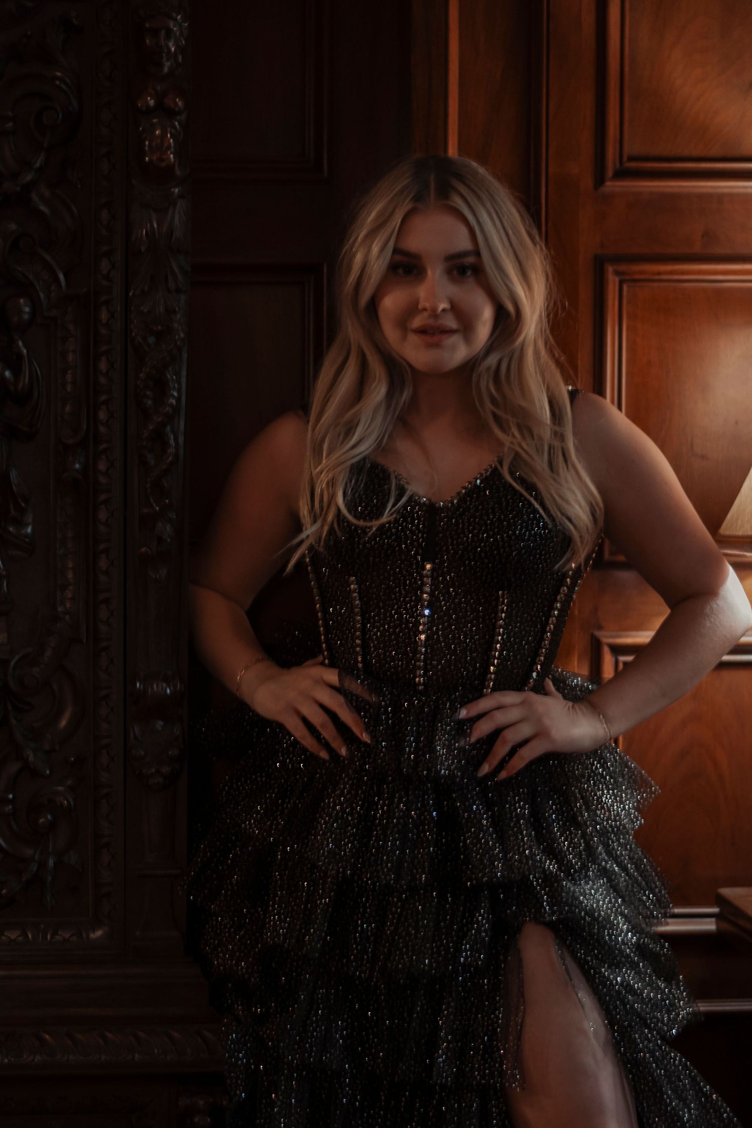Blonde woman in black sparkly corset tiered gown posing confidently by ornate carved wooden door.