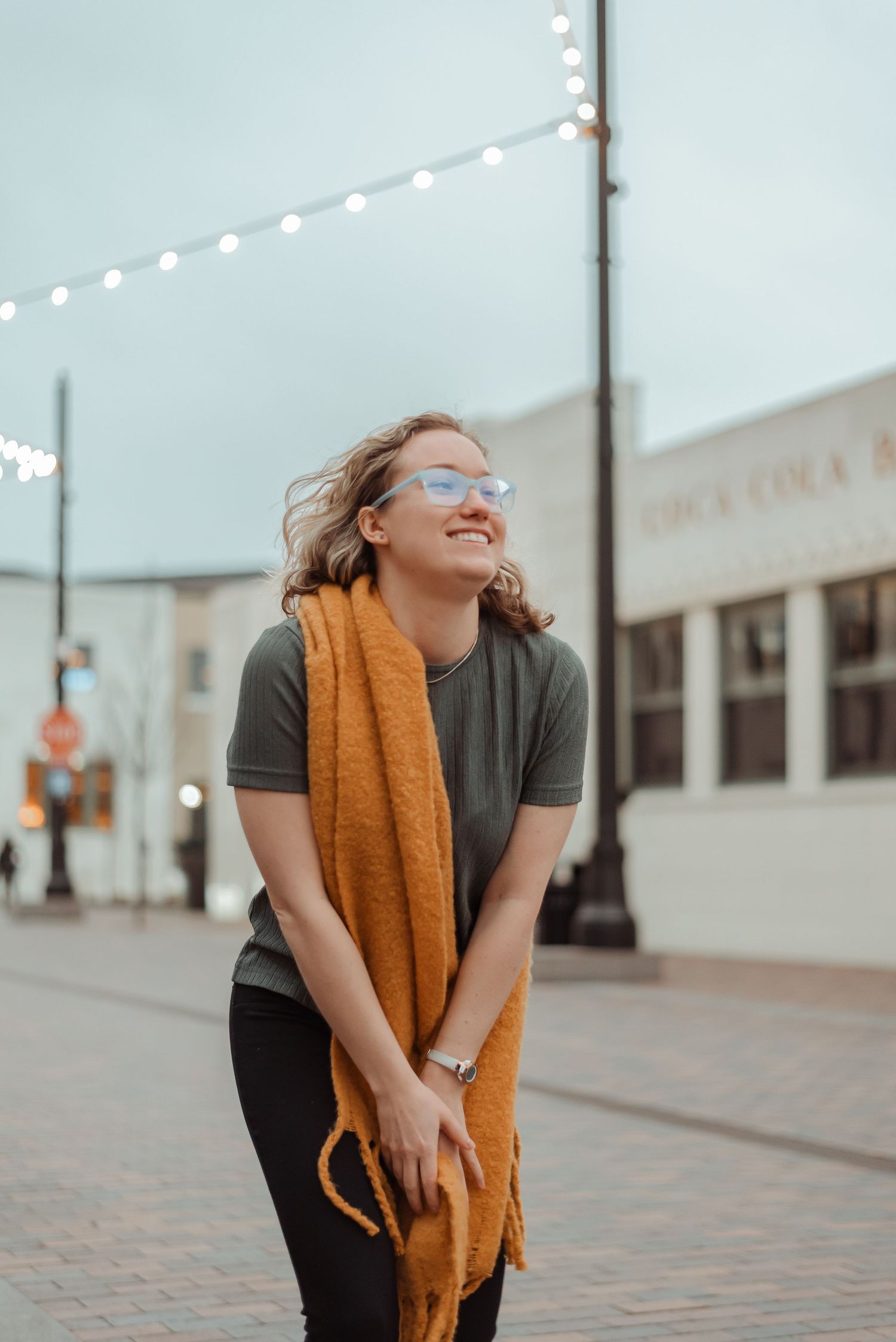Smiling young woman with glasses and mustard scarf laughing outdoors on a brick street at dusk.