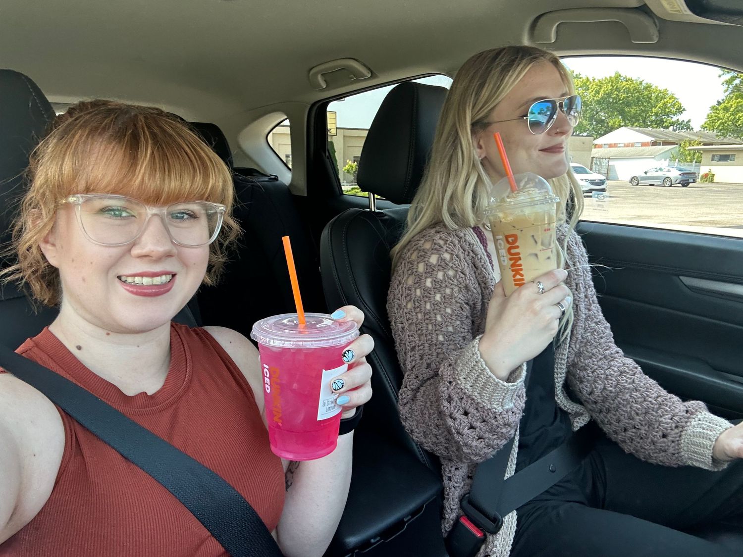 Two young women smiling in a car holding colorful drinks from a fast food restaurant.