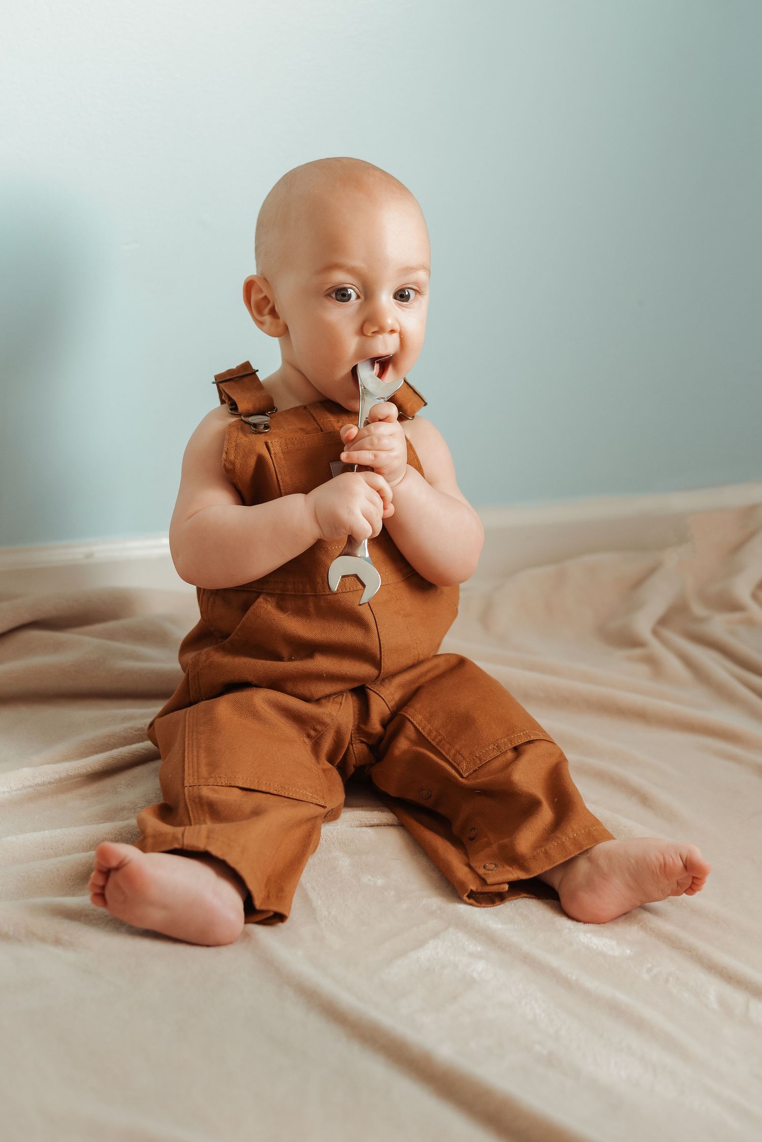 Baby in brown overalls sits on beige fabric chewing on a toy wrench.