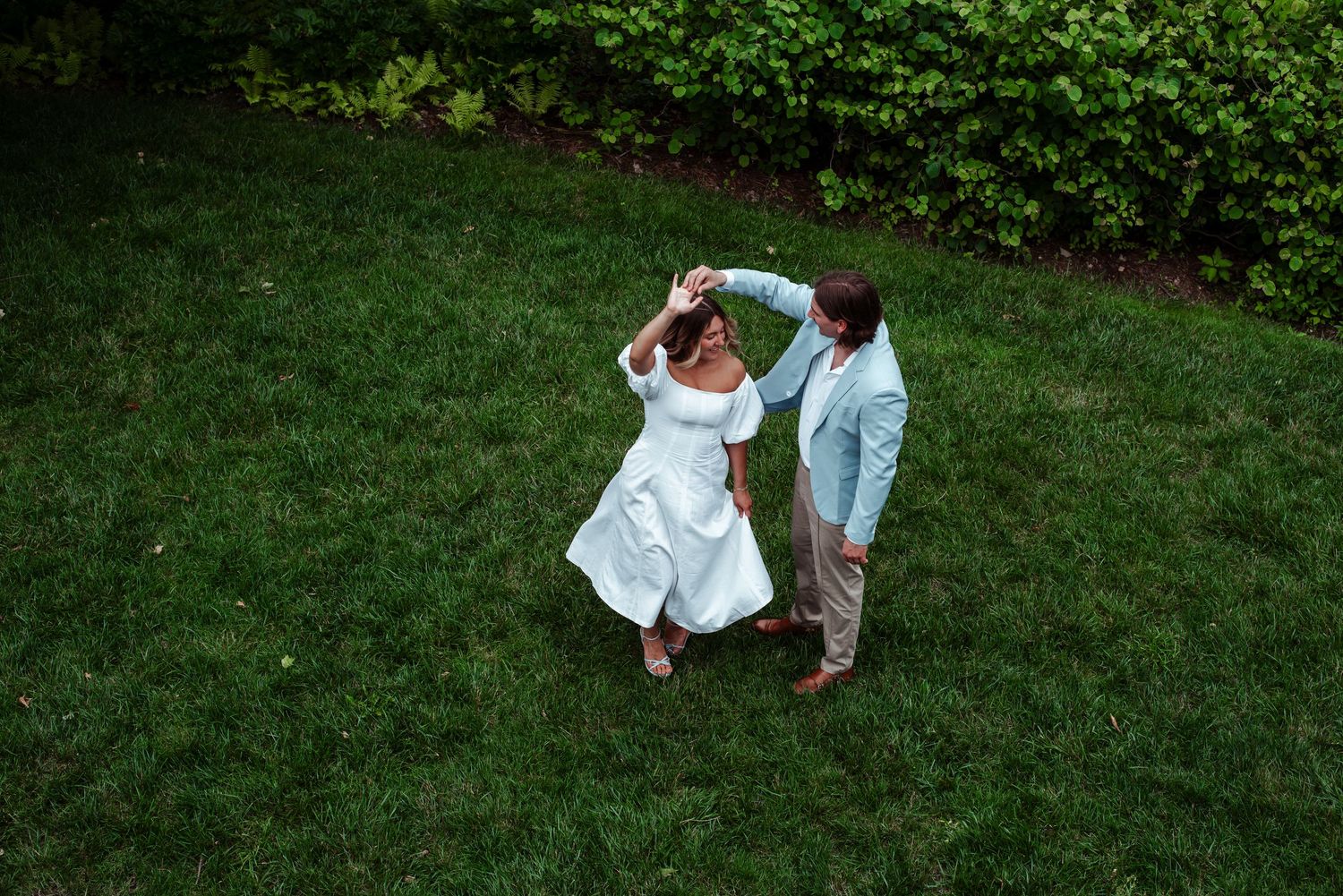 Aerial view of a bride and groom dancing on green grass, groom spinning bride in white dress and light blue suit.
