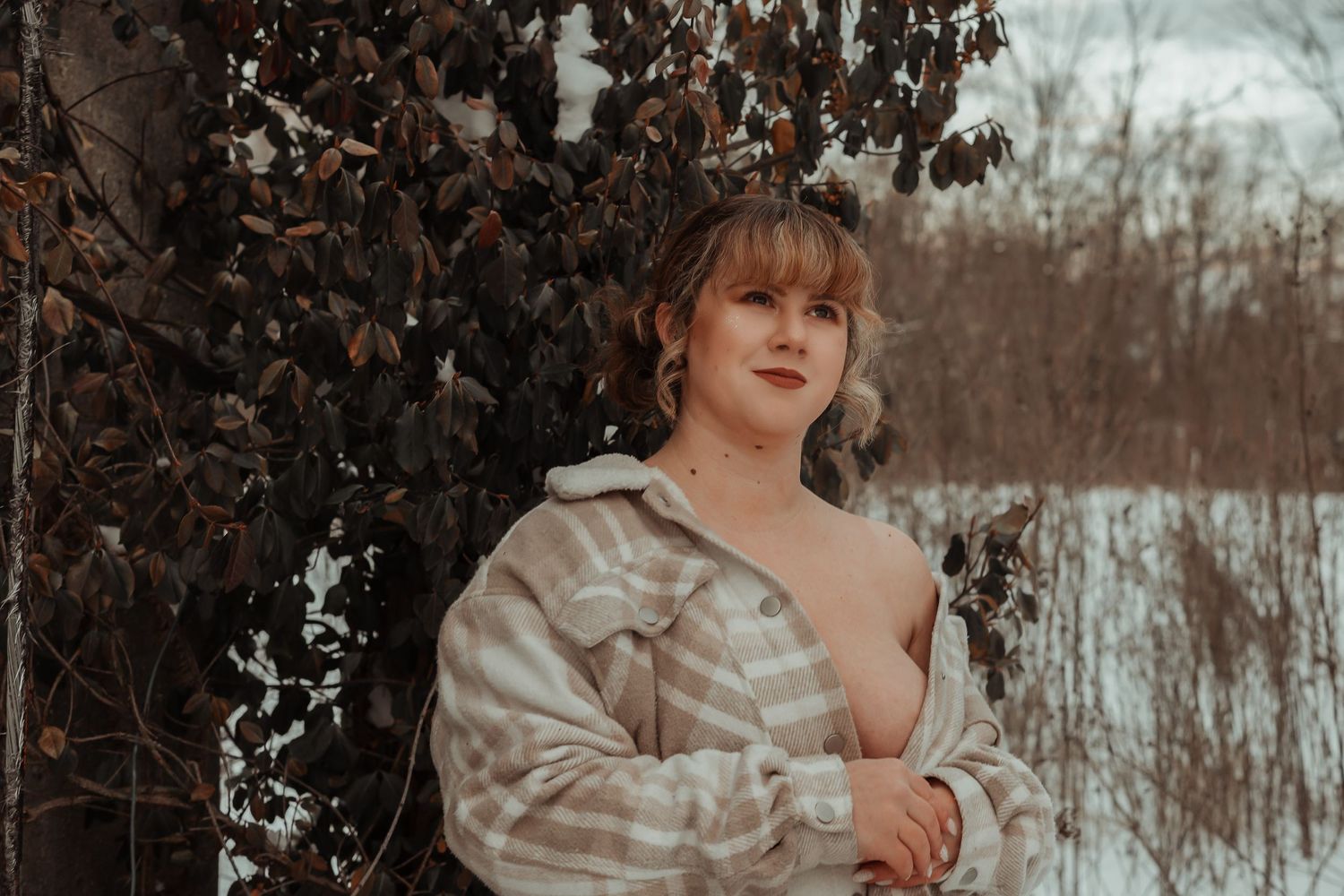 Young woman in plaid flannel shirt poses outdoors near autumn foliage with snowy background.