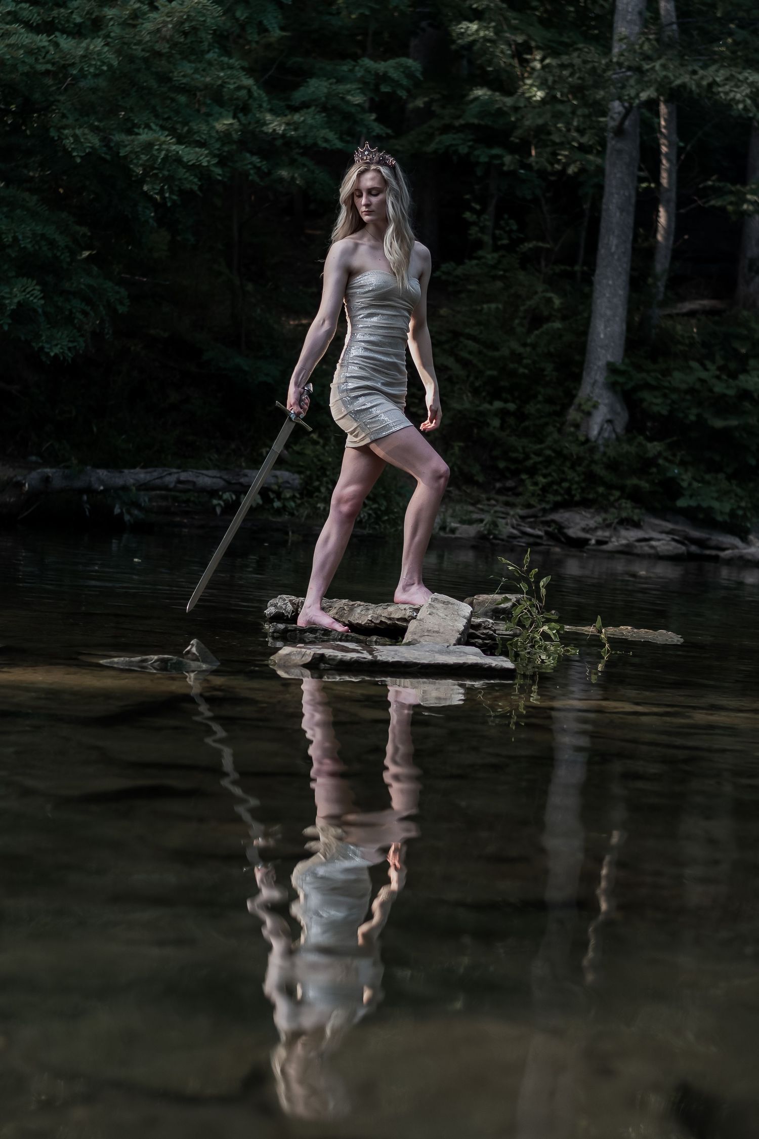 Fantasy warrior princess in silver dress and crown stands on rocks in forest stream with sword.