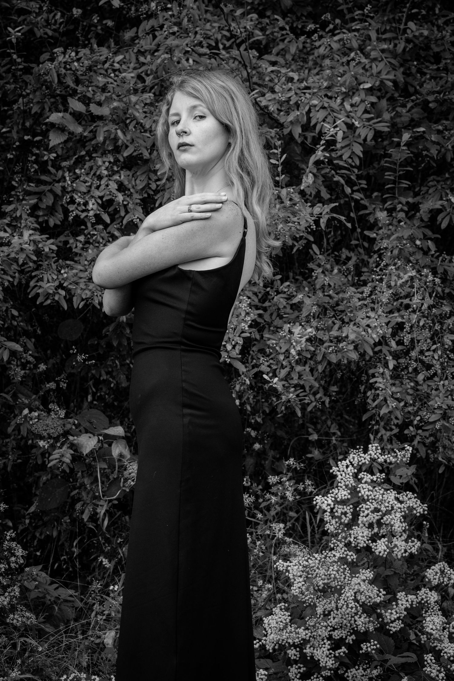 Black and white portrait of a blonde woman in a black dress with arms crossed, standing before lush foliage.