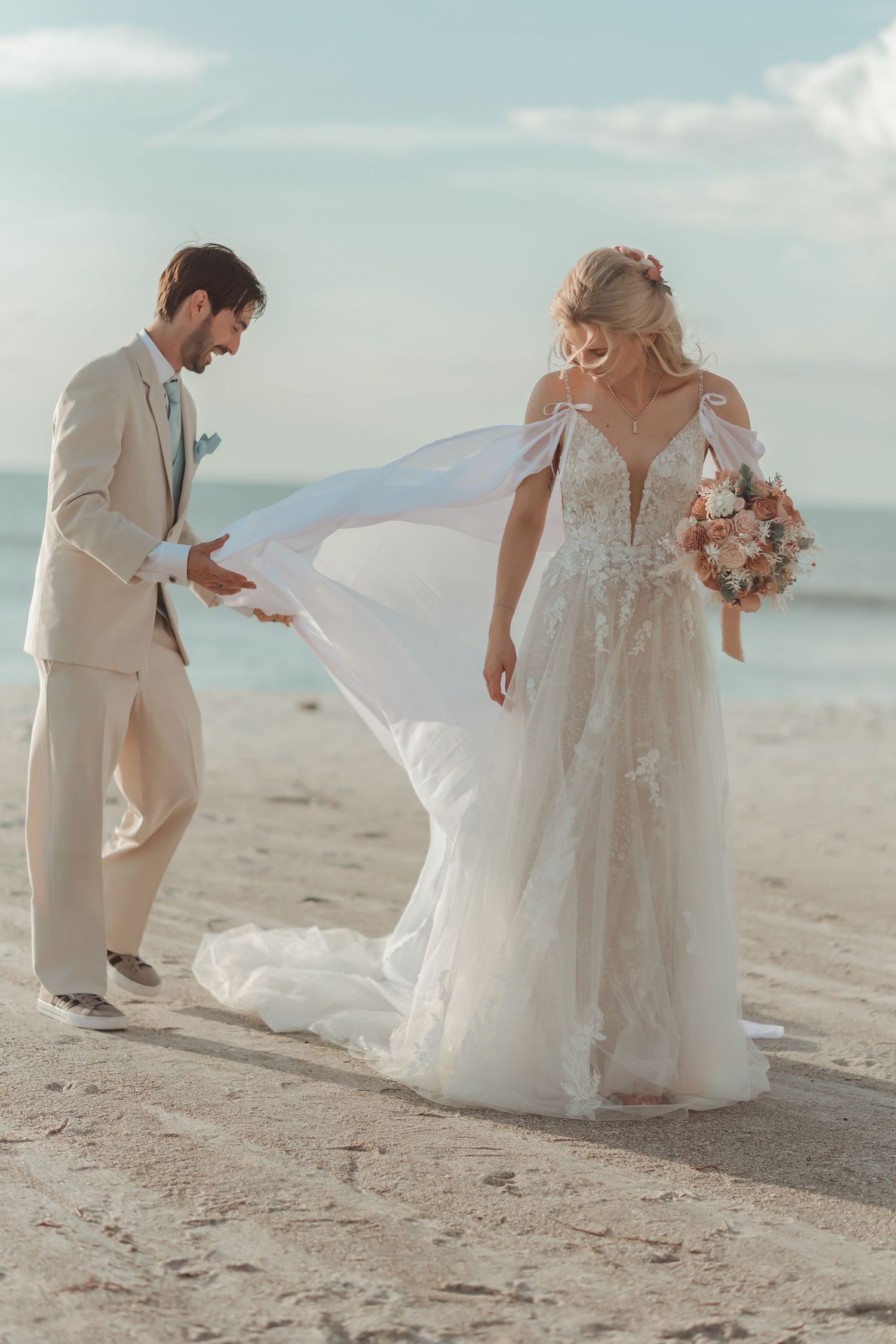 Groom in beige suit adjusting bride's flowing white lace wedding gown on sandy beach.