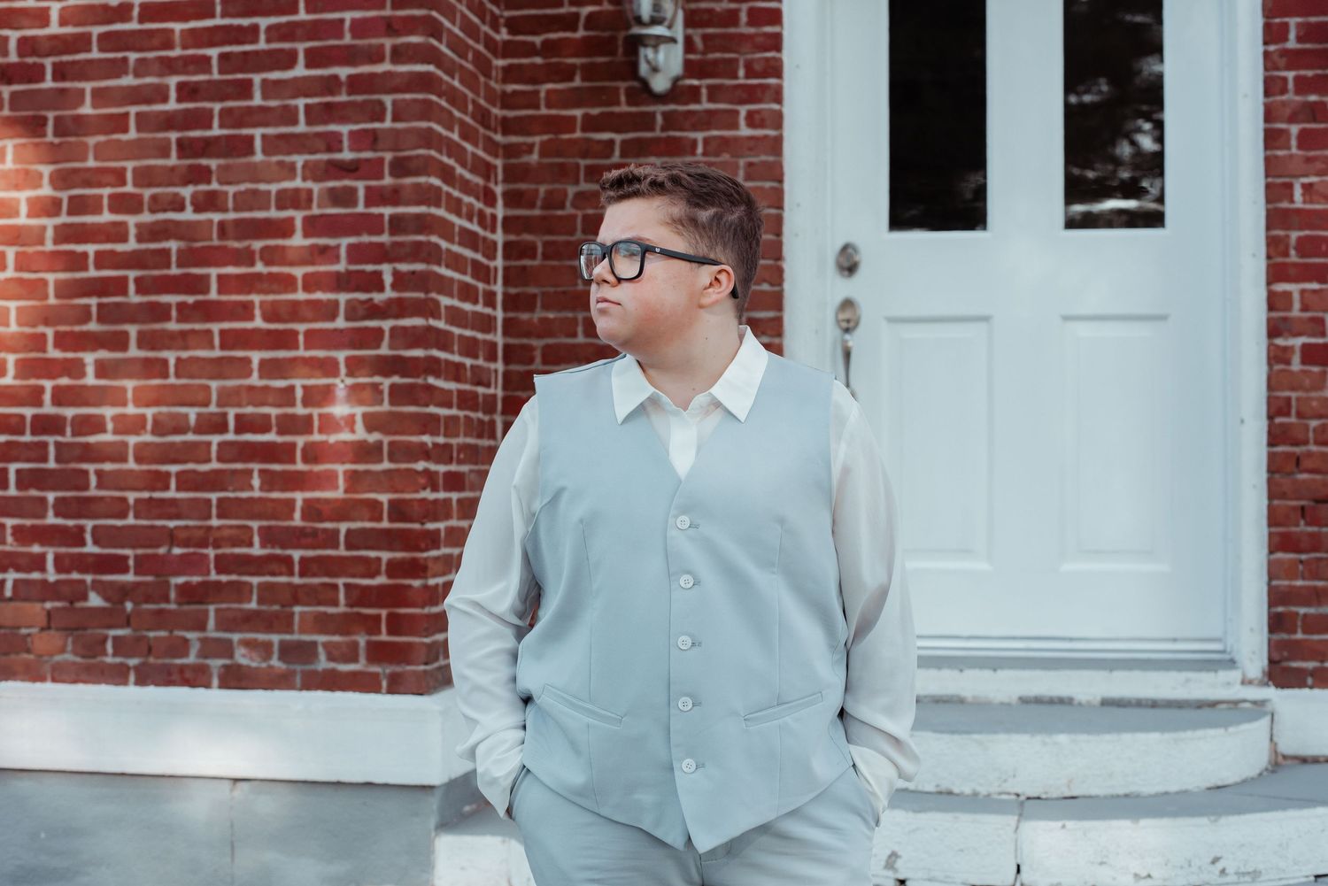 Young man in light gray vest and dress shirt poses by brick building before prom.