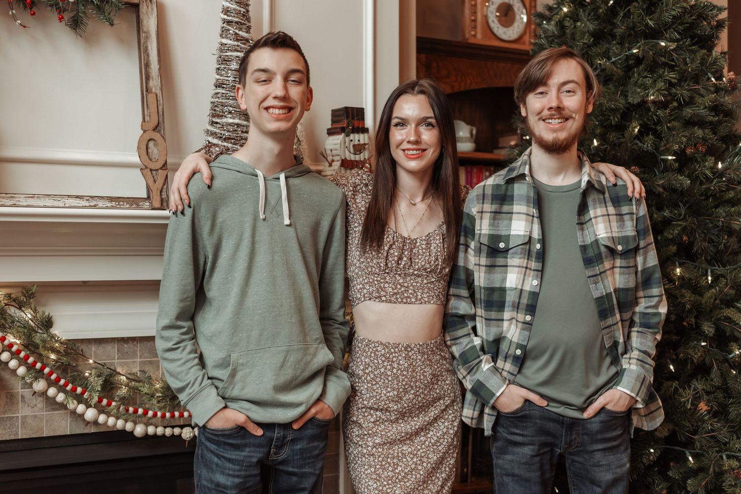 Three young adult siblings smile together indoors near a decorated Christmas tree during holiday photos.