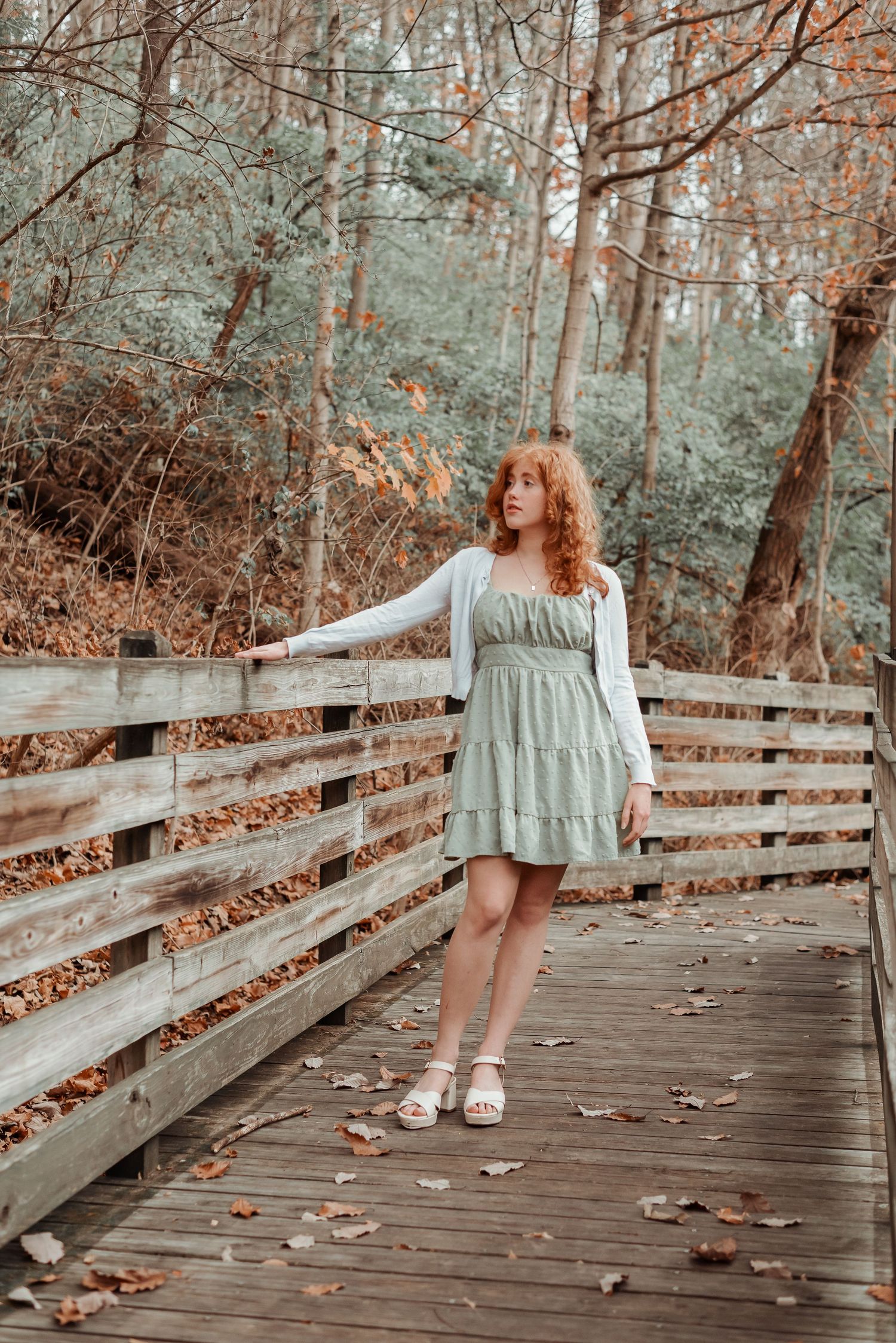 Redhead woman in sage green dress stands on wooden bridge in autumn forest.