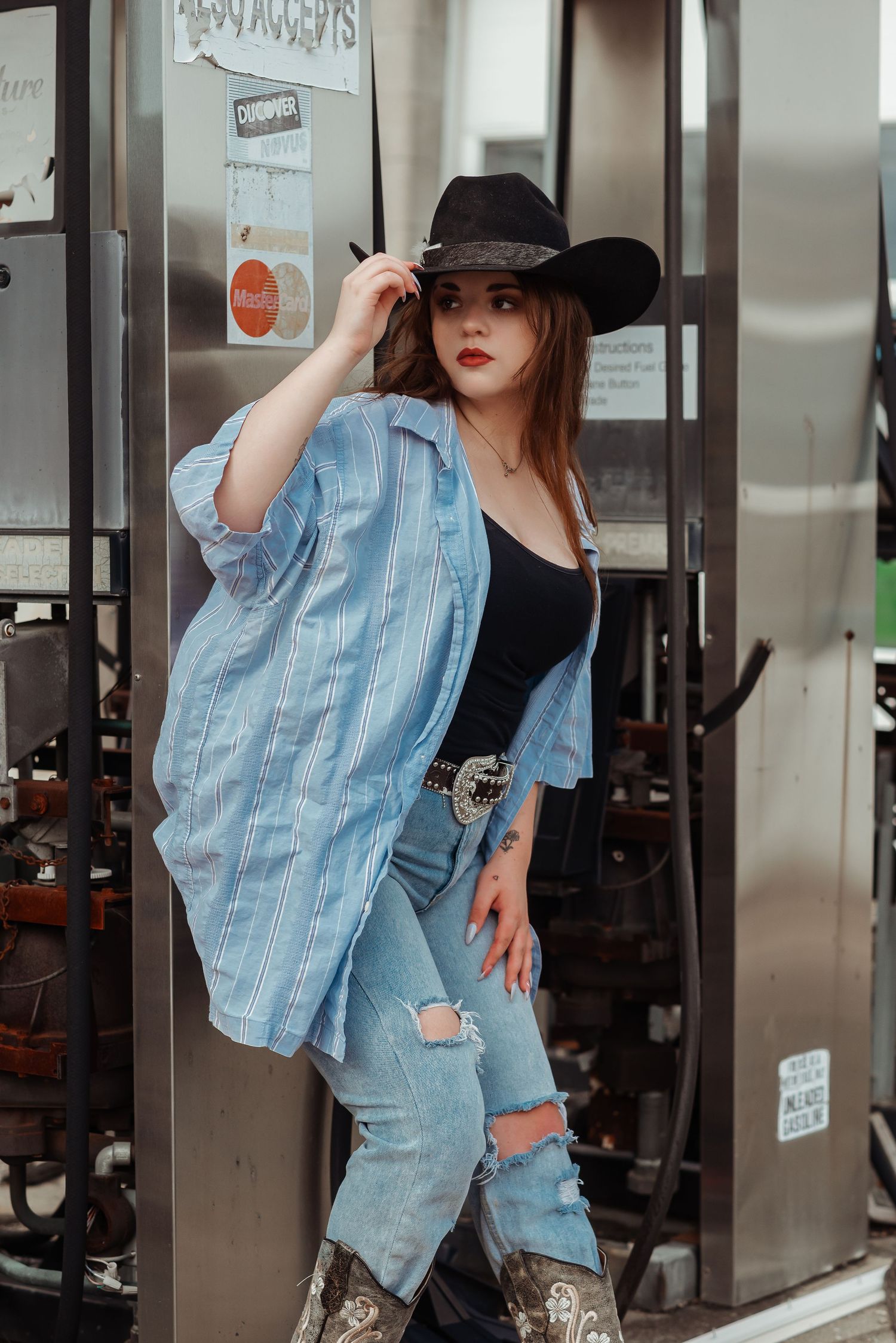 Young woman in cowboy hat, blue striped shirt, and ripped jeans poses at vintage phone booth.