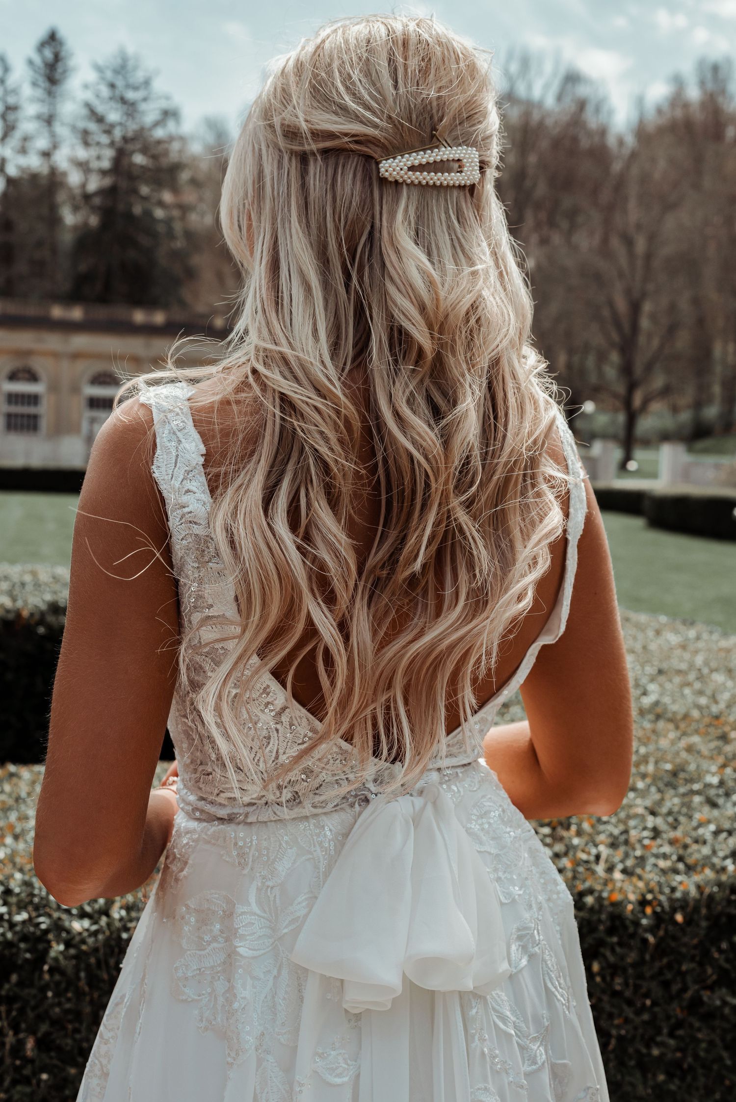 Bride with long blonde wavy hair adorned with pearl clip wearing backless lace wedding gown outdoors.