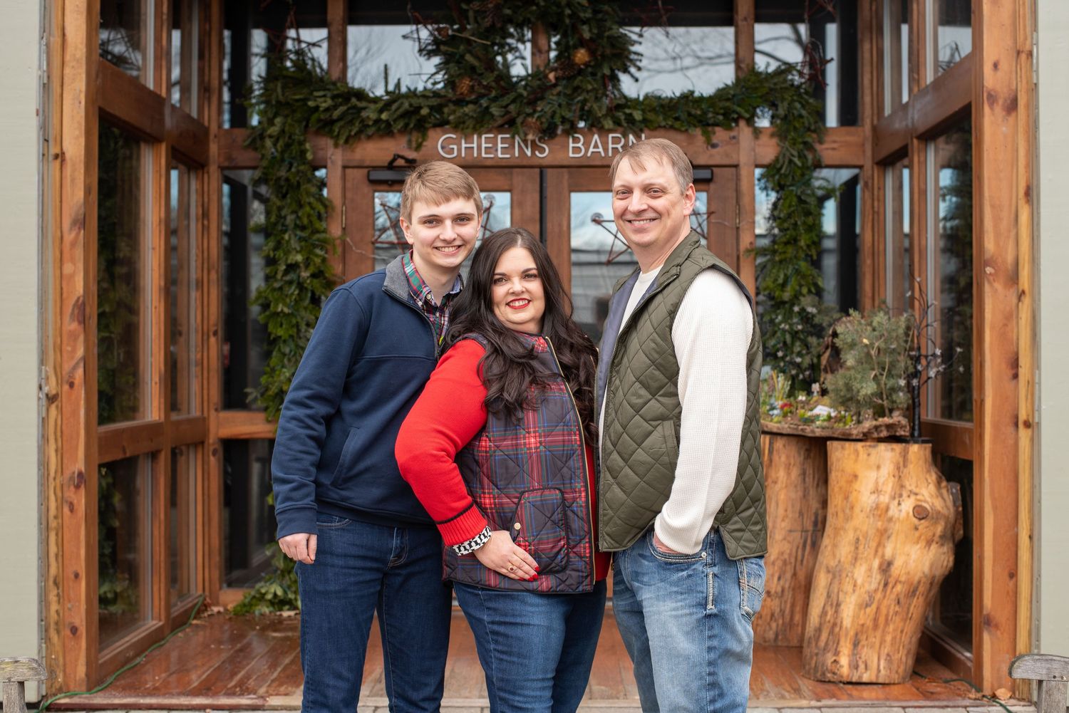 Cheerful family of three smiles together outside the Greens Barn entrance during a holiday photo session.