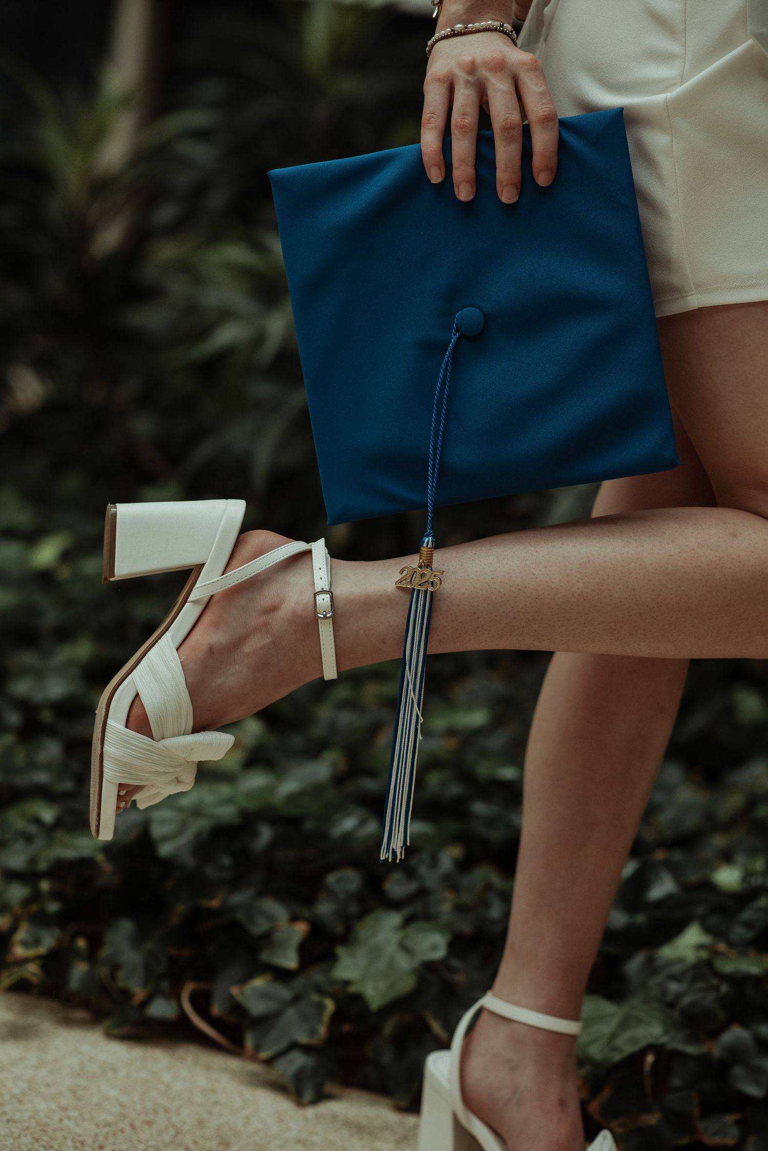 Close-up of graduate holding blue mortarboard cap with tassel while wearing white platform heels outdoors.