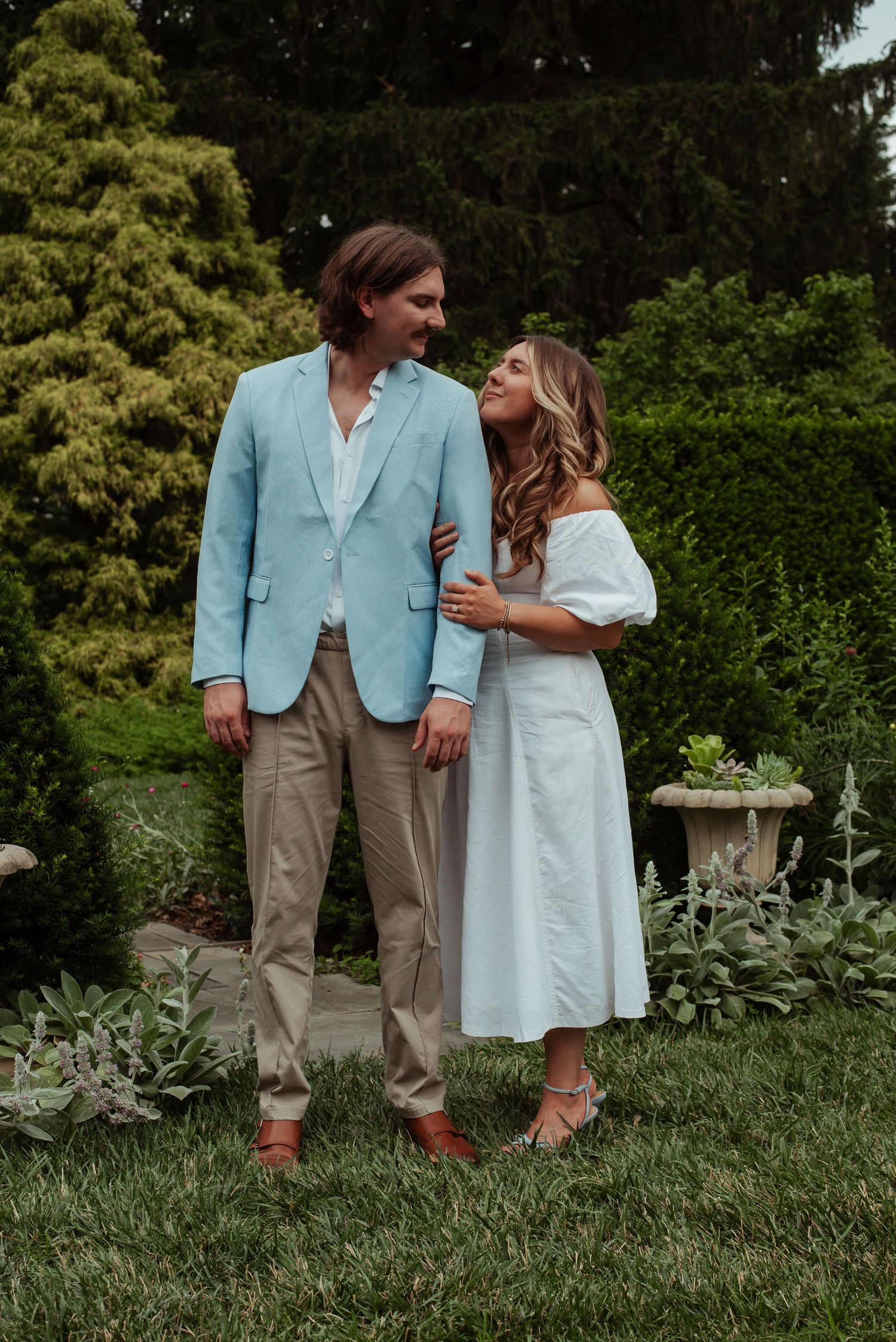 Couple in a garden, man in light blue blazer and woman in white dress, sharing a romantic moment outdoors.