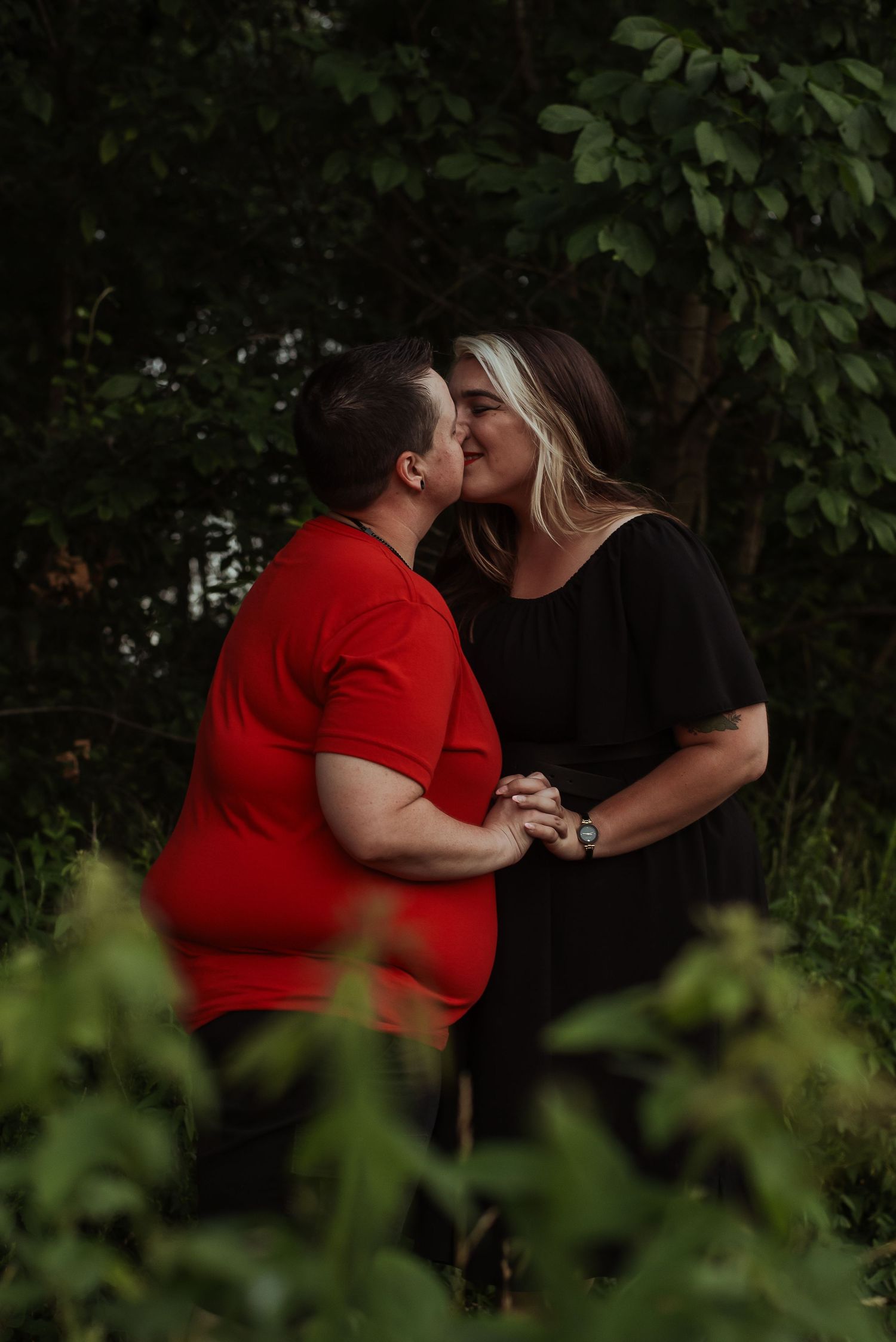 A couple sharing a romantic kiss outdoors, surrounded by lush green trees, one wearing red and the other in black.