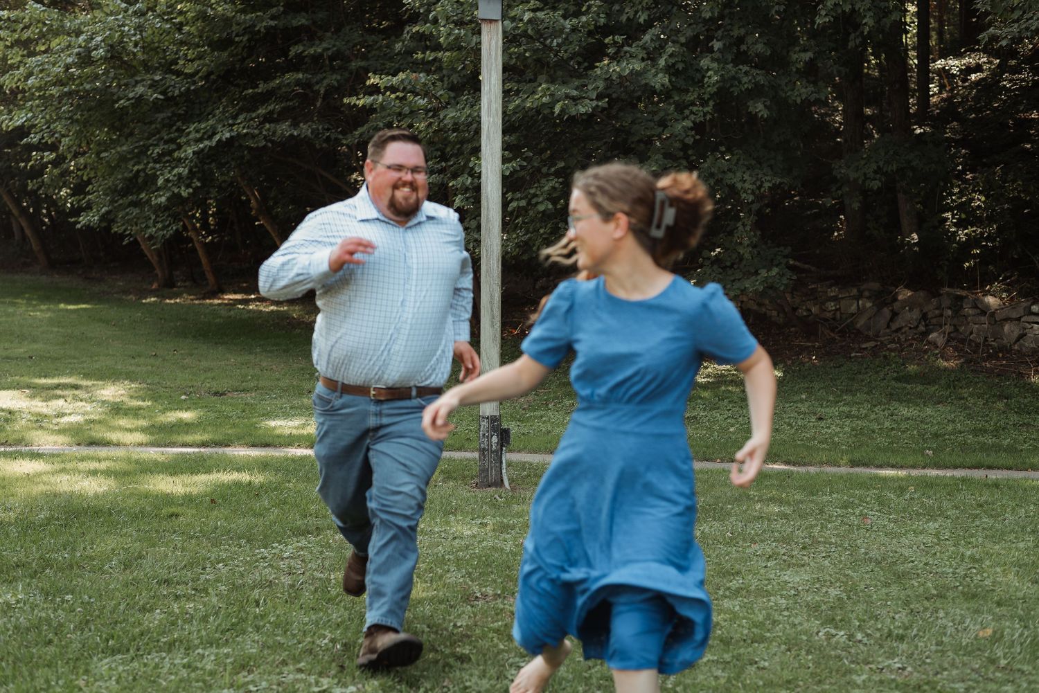 A man playfully chases a woman in a blue dress across a grassy backyard surrounded by trees.