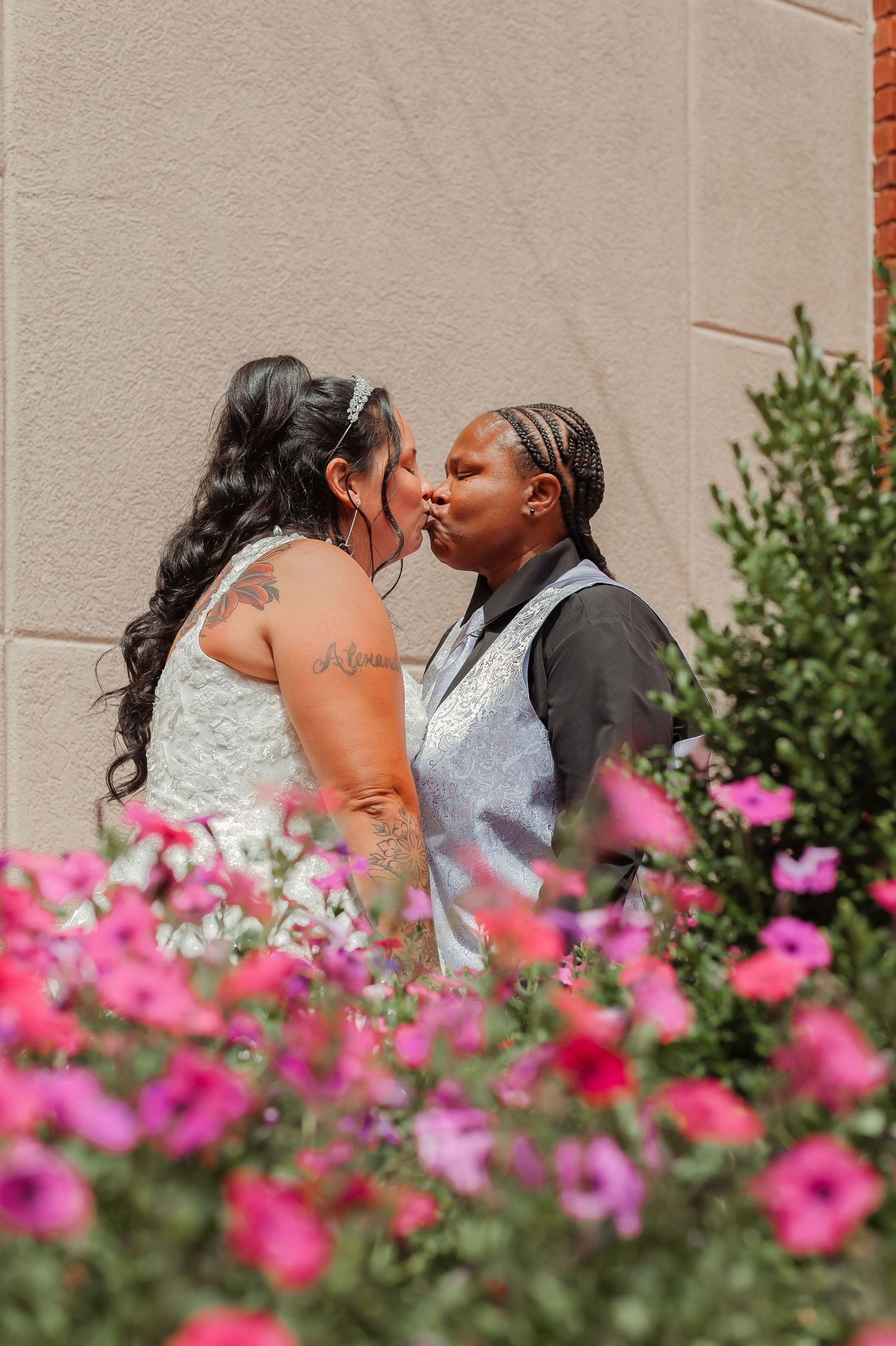 Two brides share a first kiss surrounded by vibrant pink flowers during their outdoor wedding ceremony.