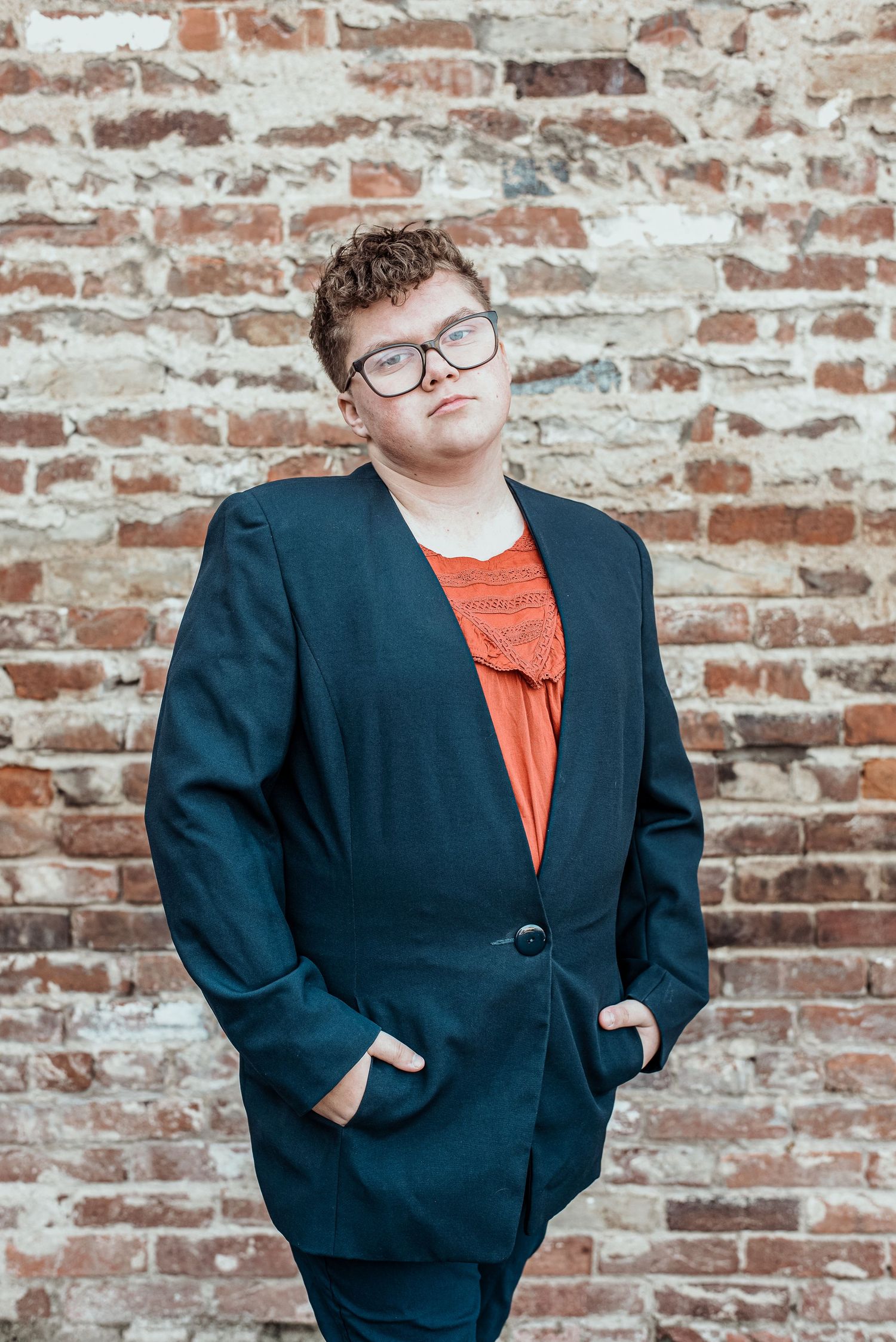 Young man in navy suit and orange shirt posing against brick wall.