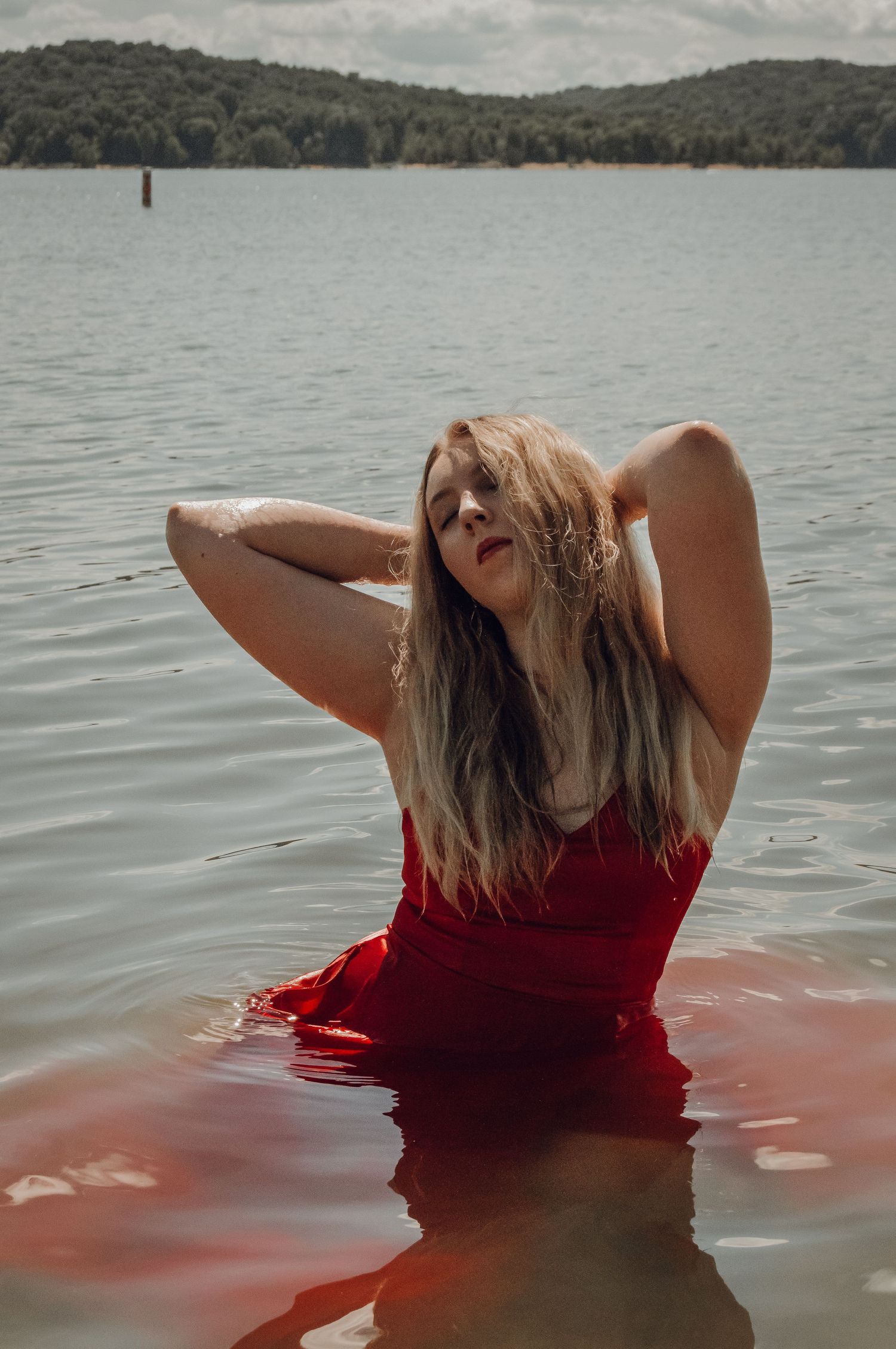Blonde woman in red dress standing in a lake with mountains in the background.