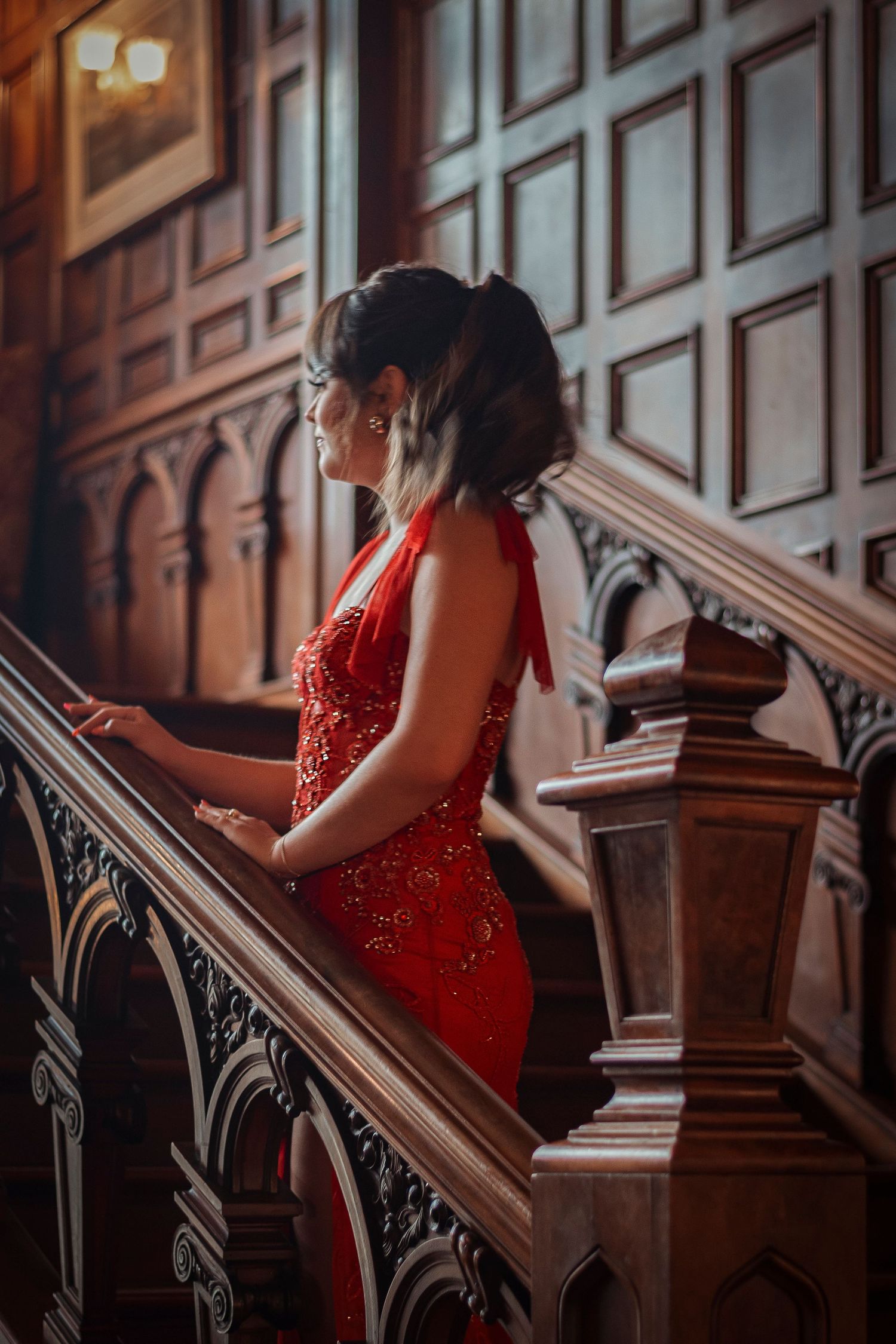 Woman in backless red sequin dress standing on carved wooden staircase in grand historic interior.
