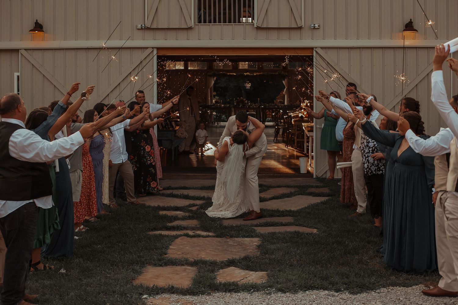 Bride and groom share a kiss during a sparkler send-off exit outside a rustic barn venue.