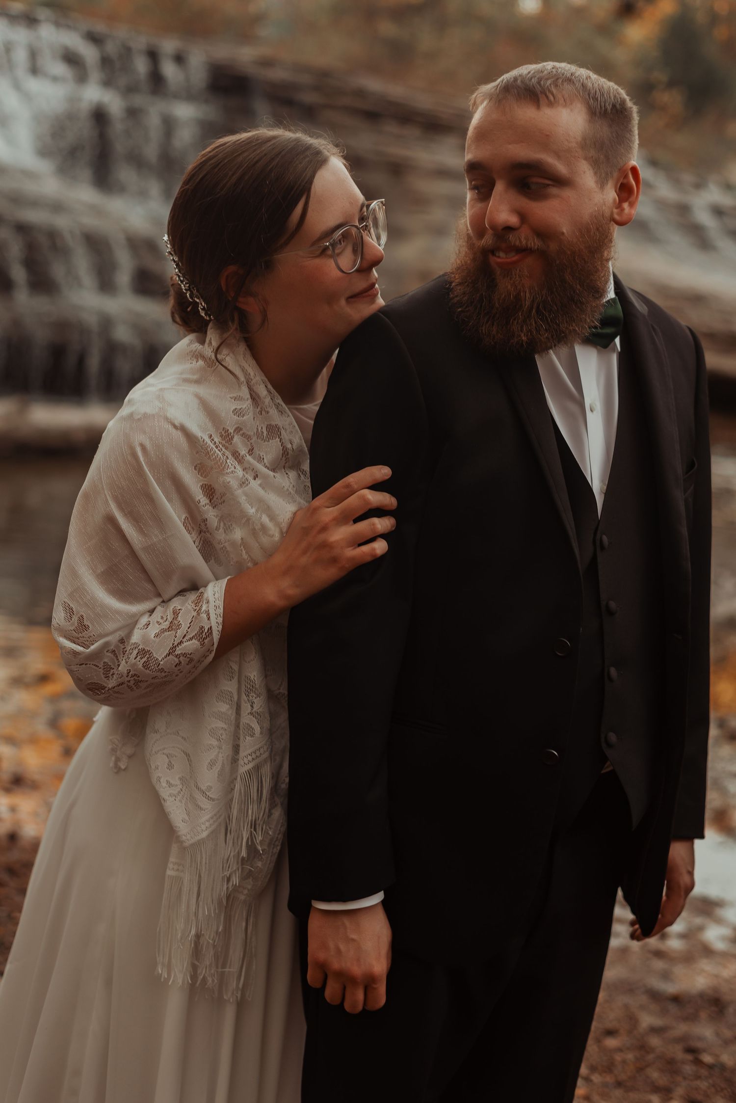 Bride in lace shawl embraces bearded groom near waterfall backdrop.