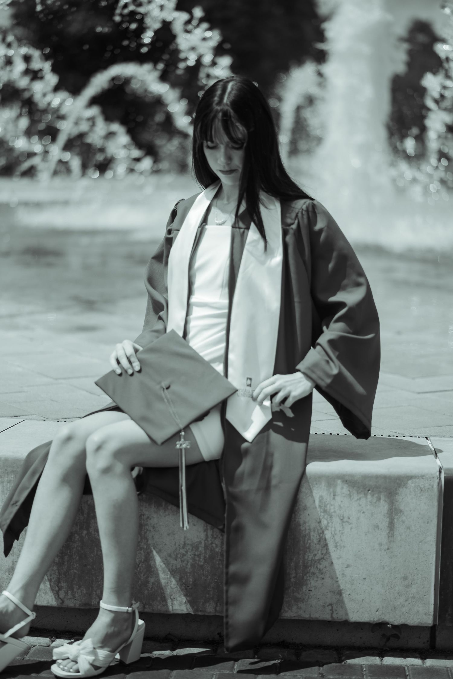 Black and white photo of graduate in cap and gown sitting by fountain holding mortarboard cap.