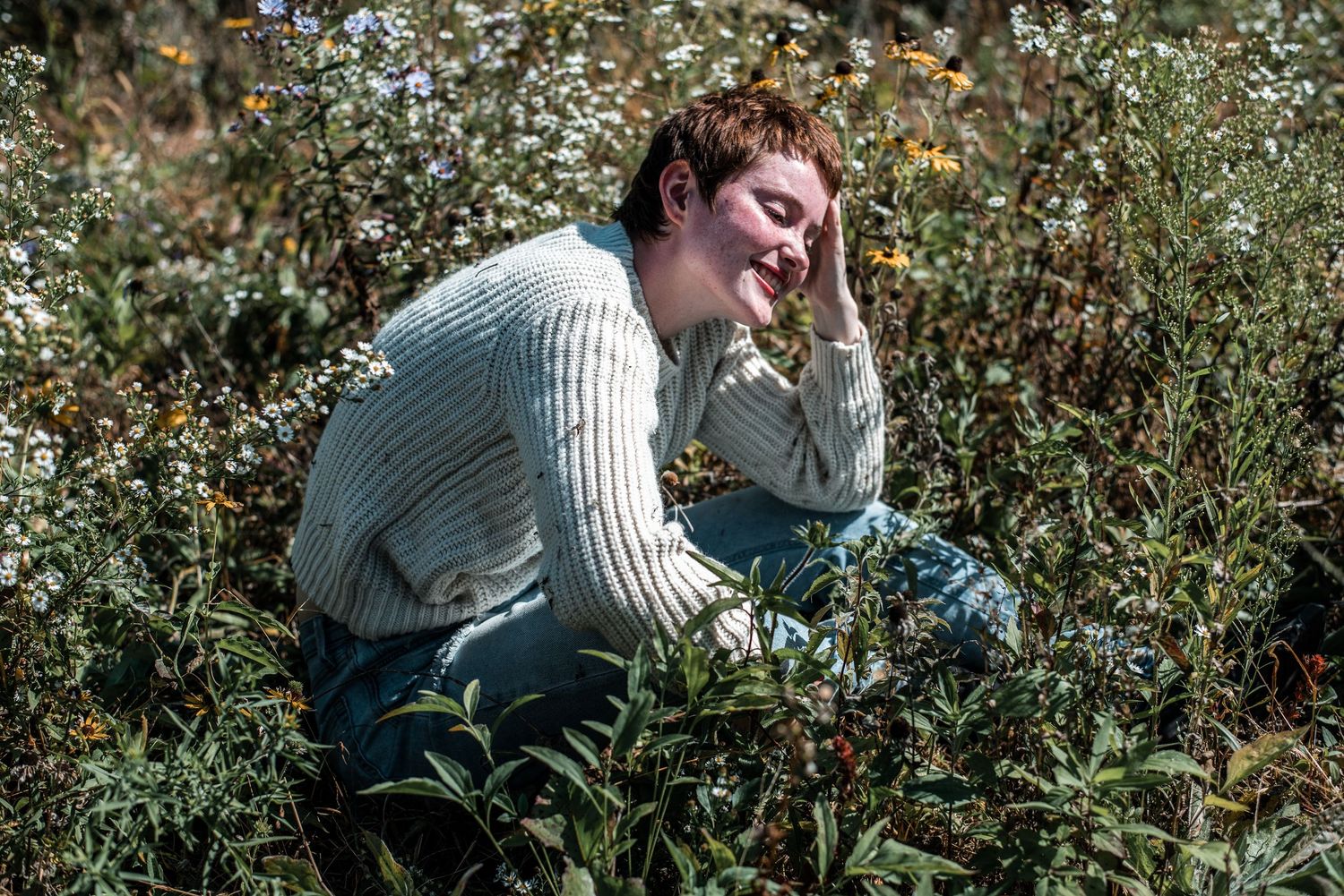A young person with short hair sits smiling among wildflowers in a sunlit outdoor field.