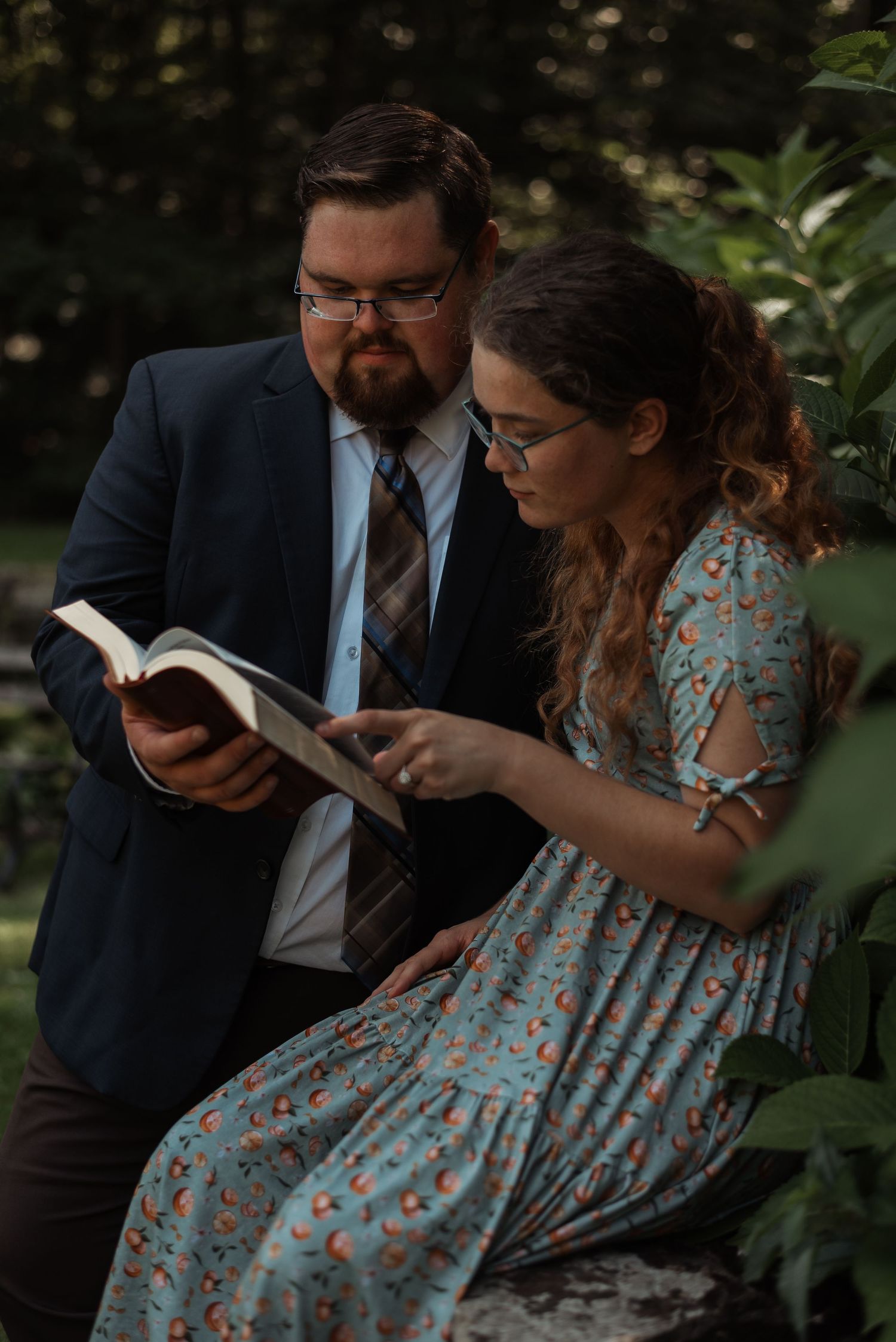 A bearded man in a suit and a woman in a floral dress read a book together outdoors.