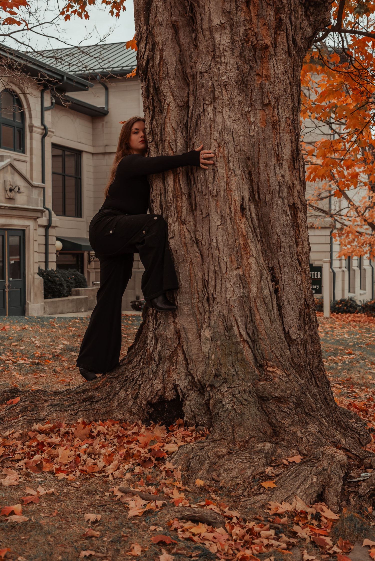 Young woman in all-black outfit embraces large tree trunk surrounded by fallen autumn leaves.